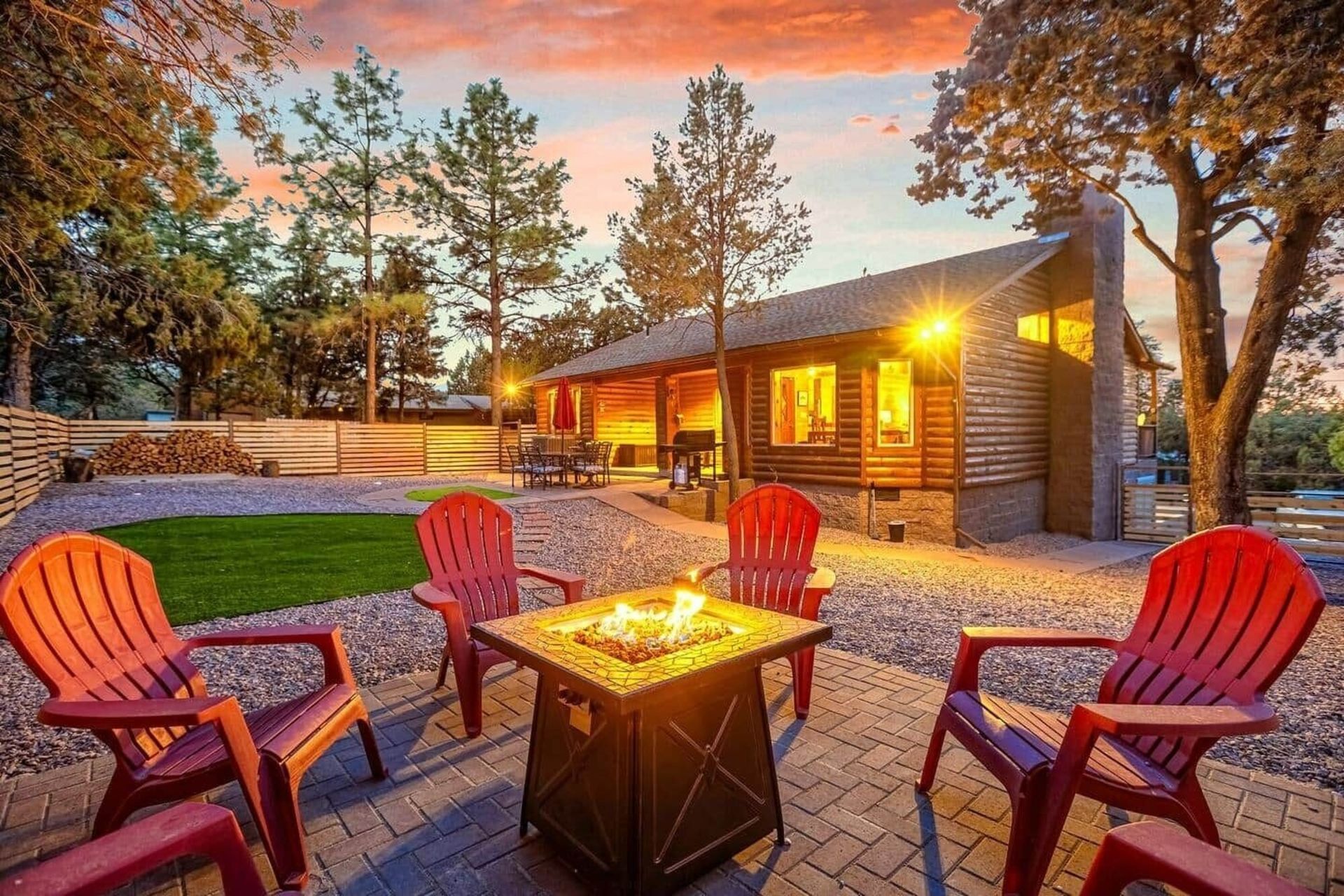 Cozy cabin patio at sunset with red chairs around a fire pit and warm lights glowing