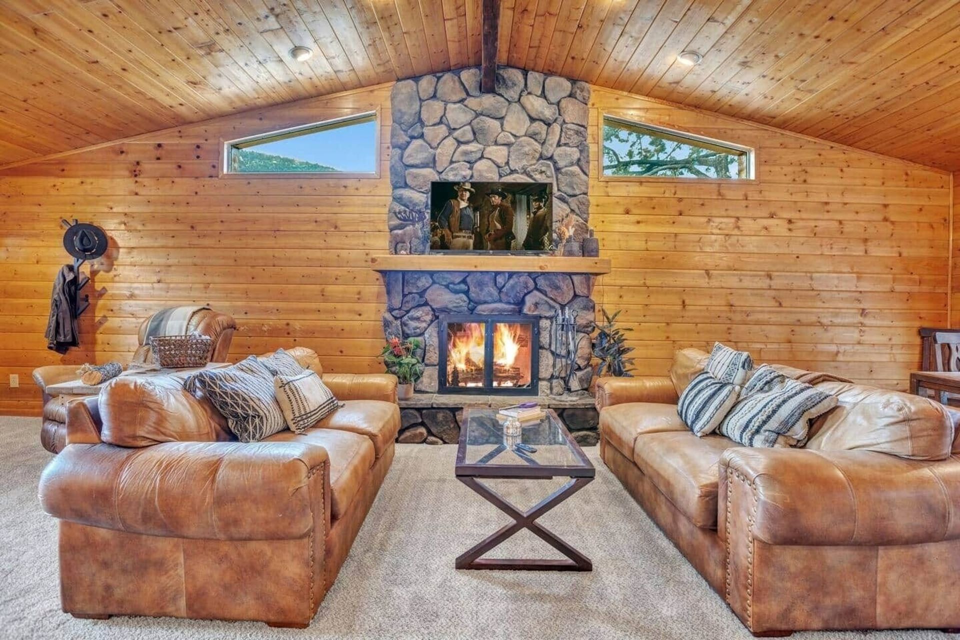 Cozy cabin living room with leather sofas, stone fireplace, and wood-paneled vaulted ceiling