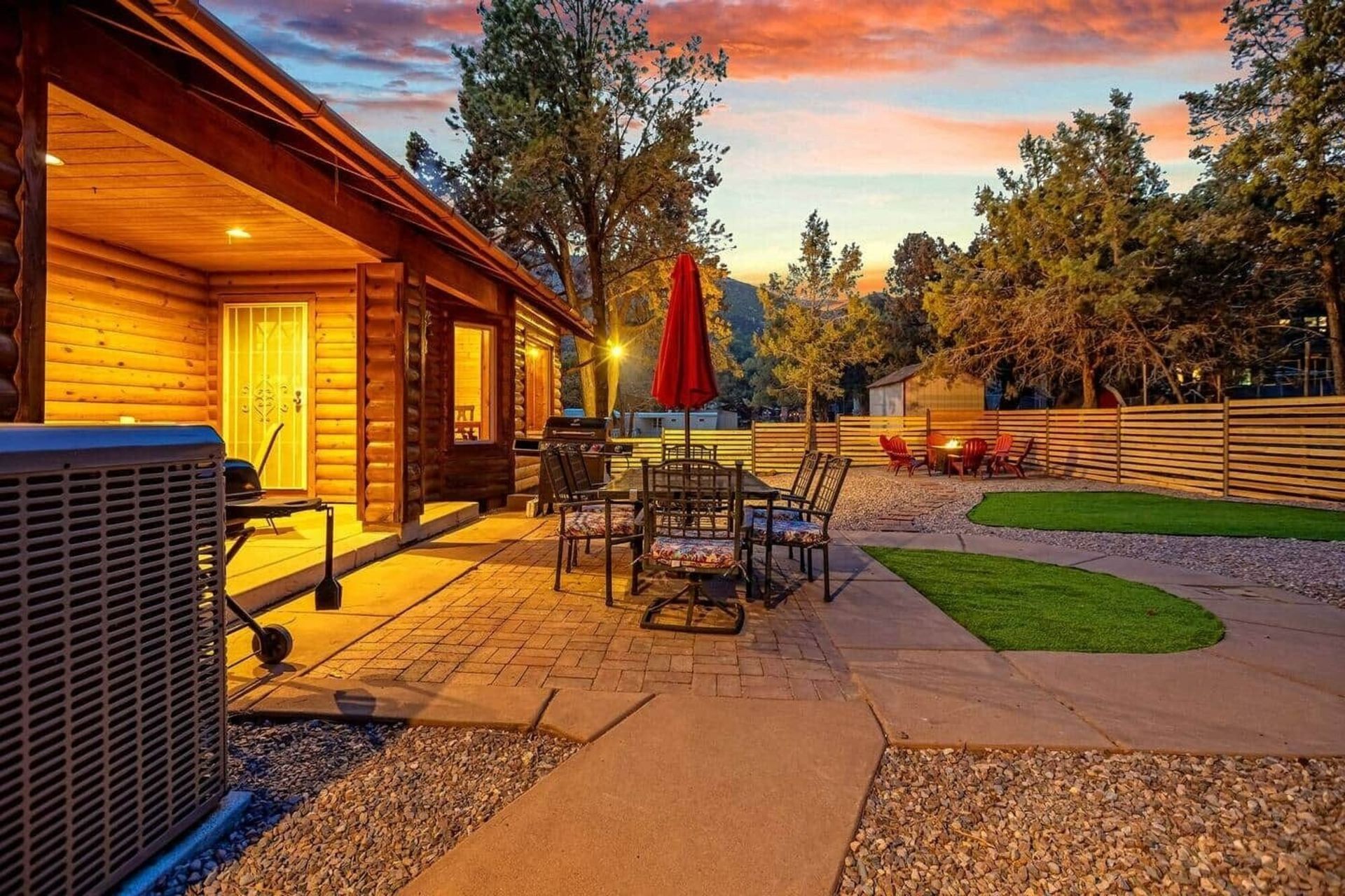 Backyard patio at sunset with outdoor dining table, red umbrella, fire pit, and lit cabin-style house
