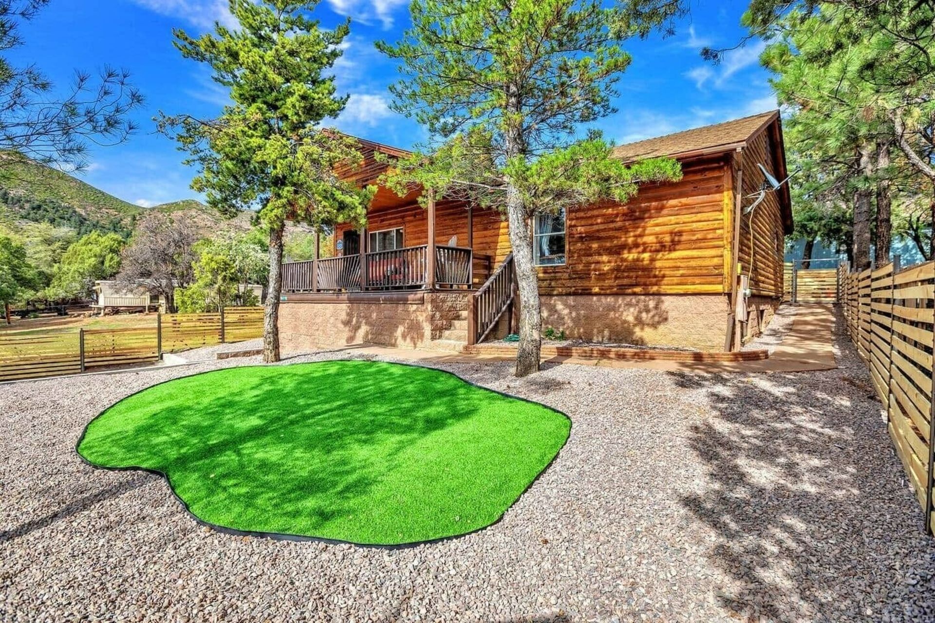 Rustic wooden cabin with porch, gravel yard, green lawn patch, and trees under a blue sky