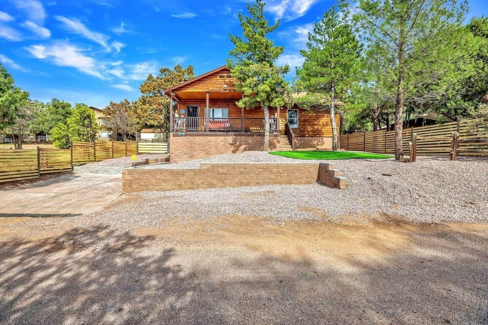 Rustic cabin with red roof and stone porch, surrounded by trees and a gravel driveway under a blue sky