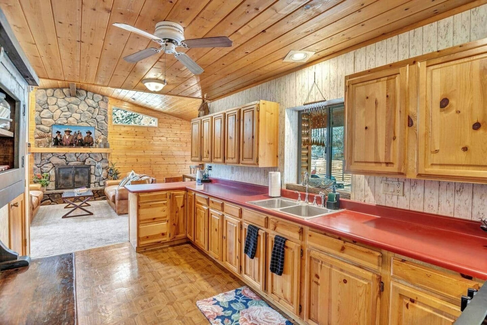 Rustic wood kitchen with red countertops, ceiling fan, and stone fireplace in the background