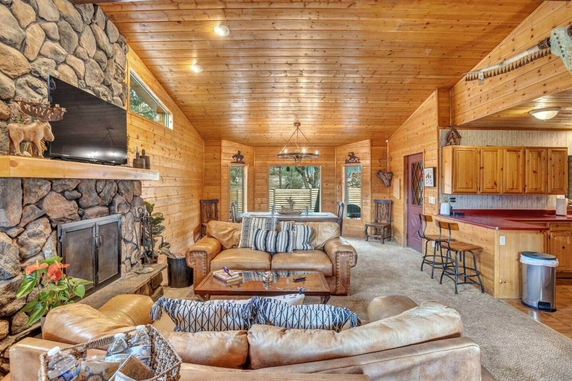 Cozy log cabin living room with stone fireplace, leather sofas, wood walls, and open kitchen area