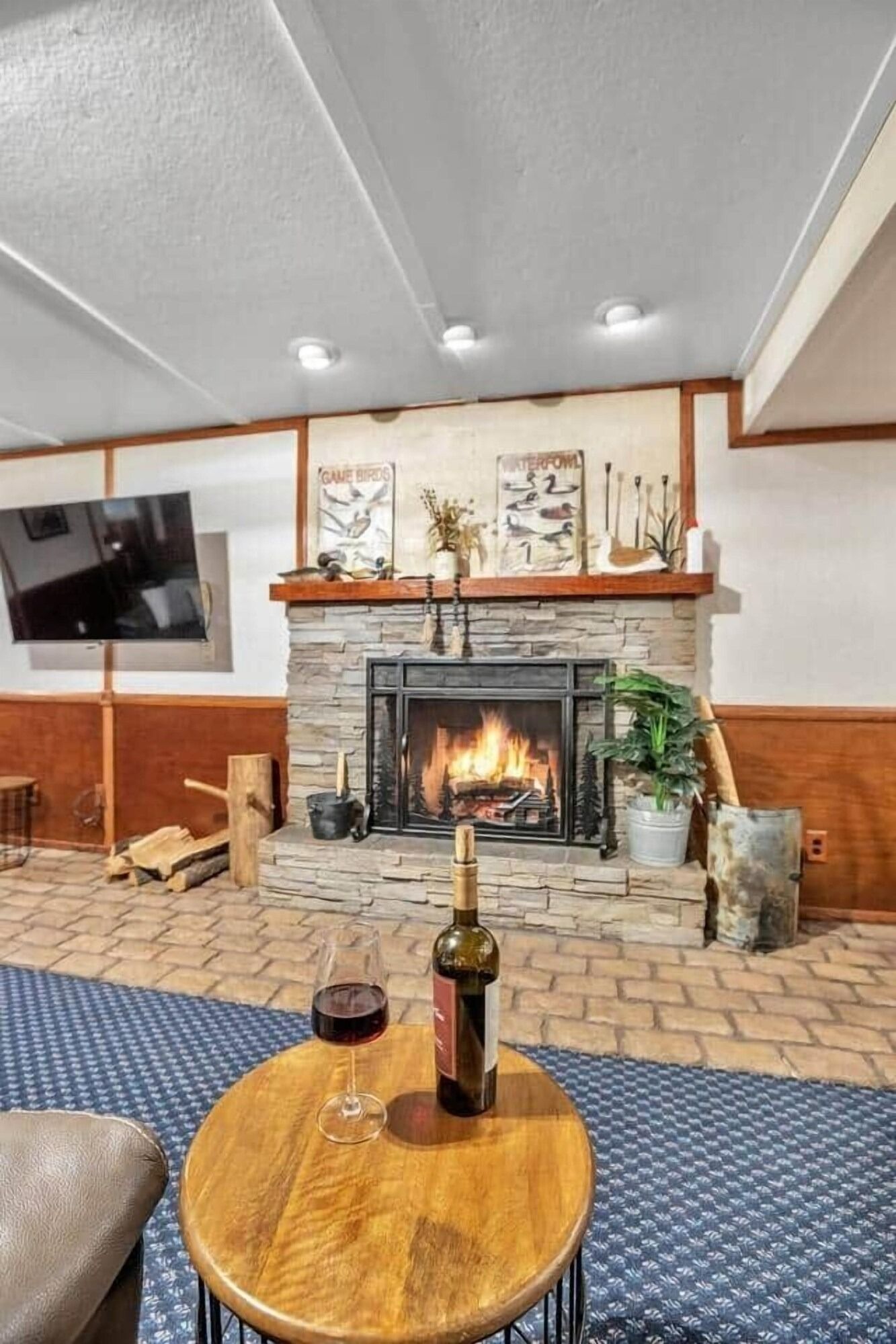 Cozy lounge with lit stone fireplace, wood paneling, blue carpet, and a wine bottle on a round table