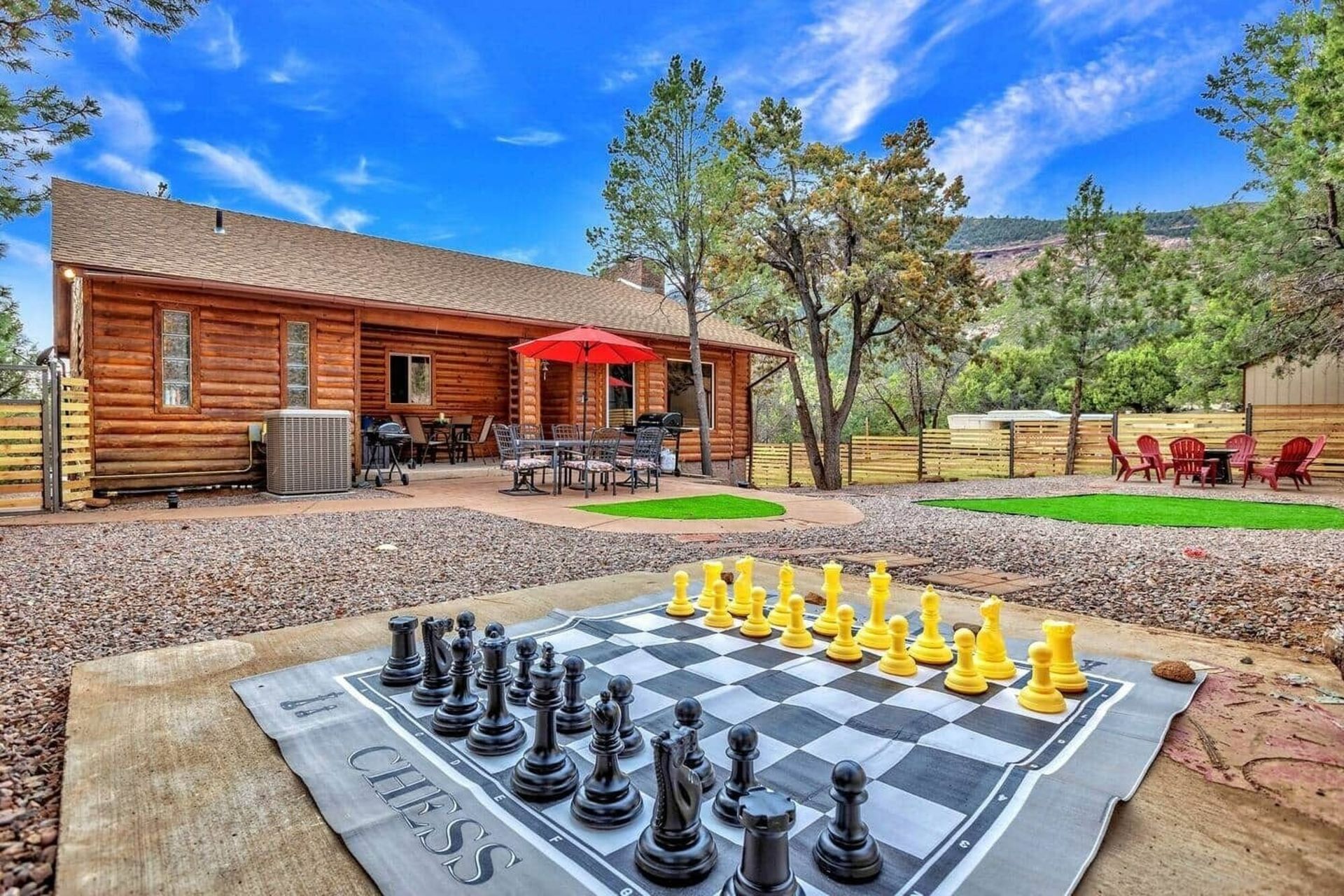 Large outdoor chessboard on a patio near a log cabin with red chairs and trees under a blue sky