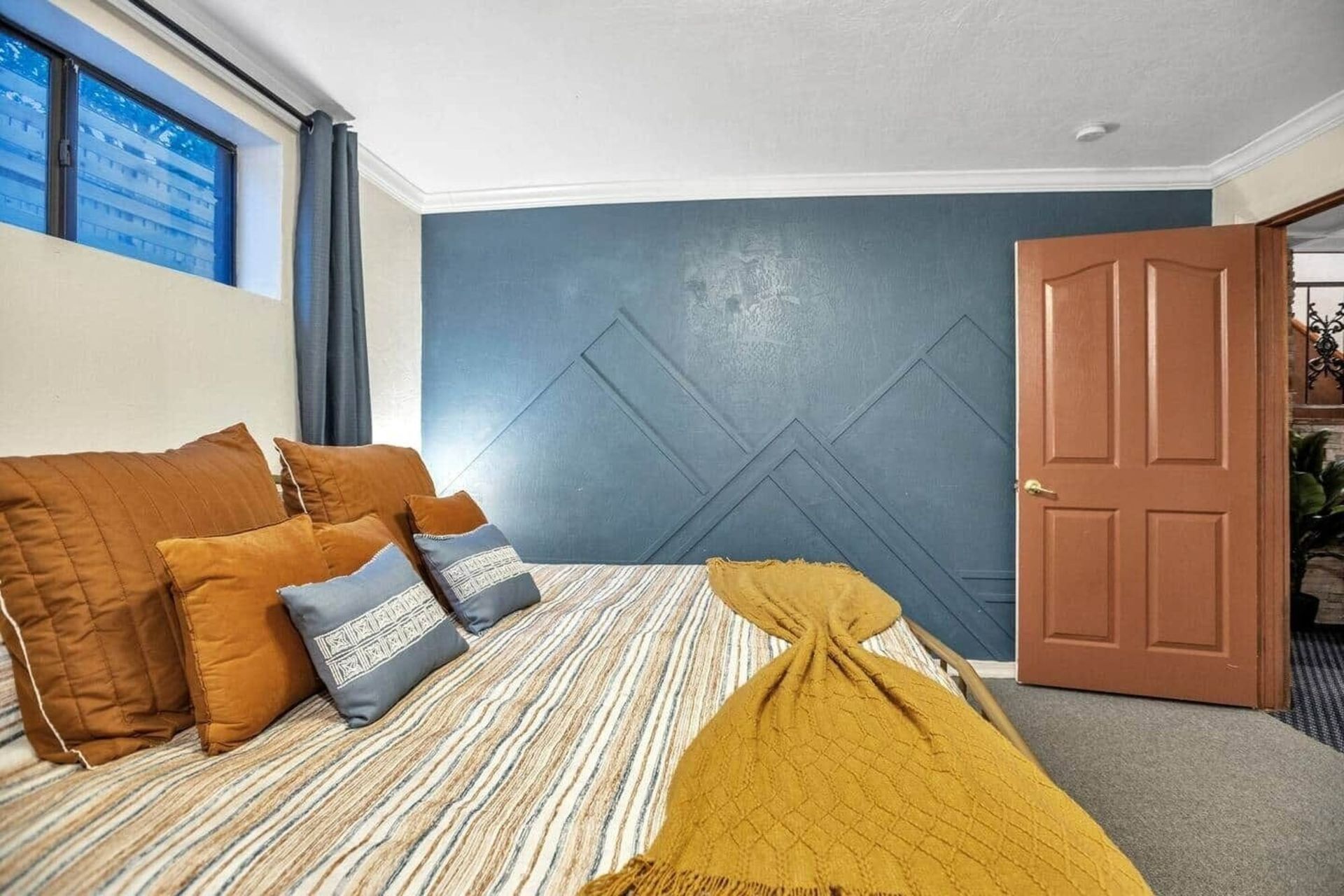 Bedroom with blue walls, mustard bedding, and an open wooden door beside a window.