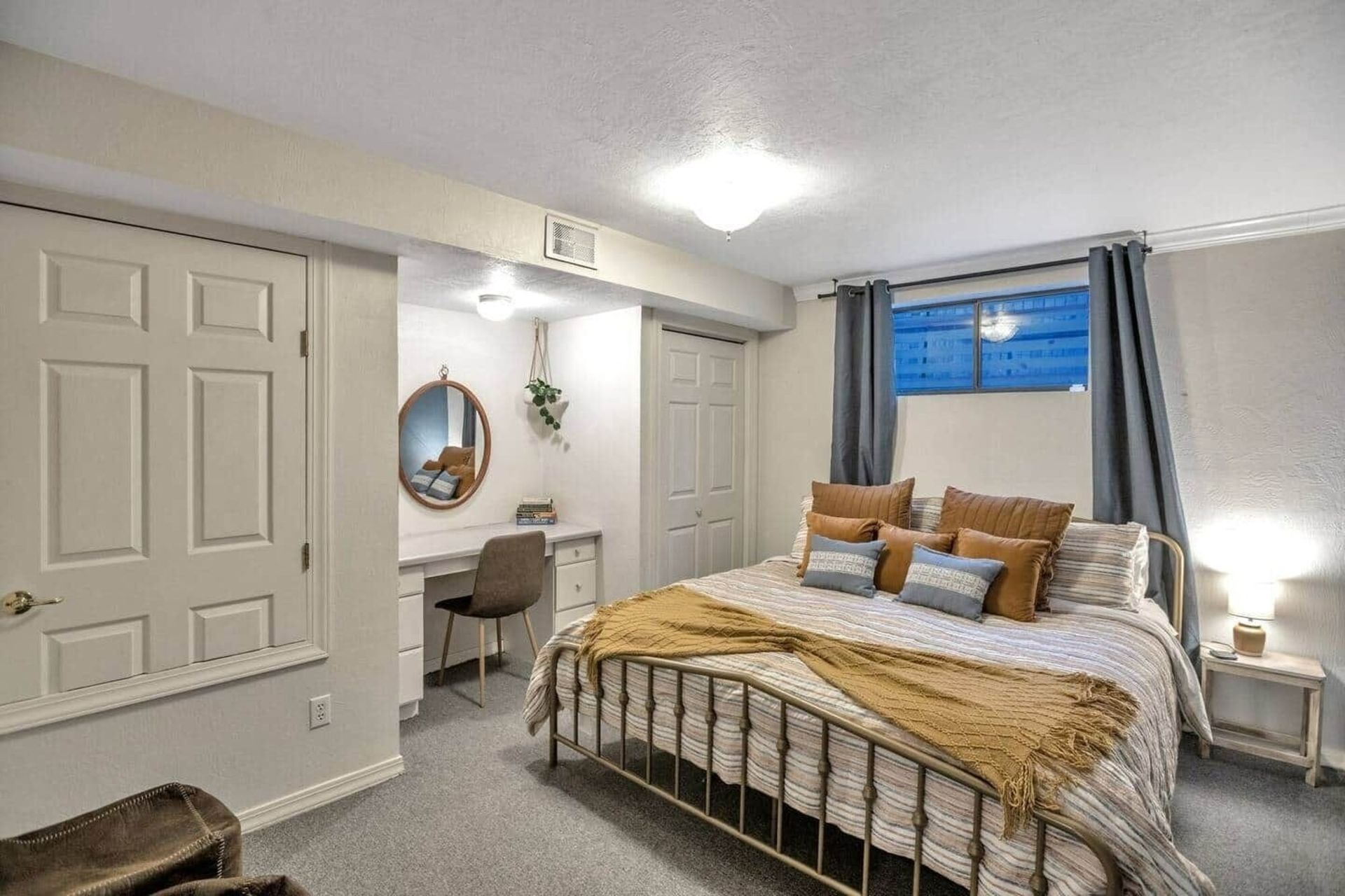 Small bedroom with a metal bed, vanity desk, beige walls, and blue curtains by the window.