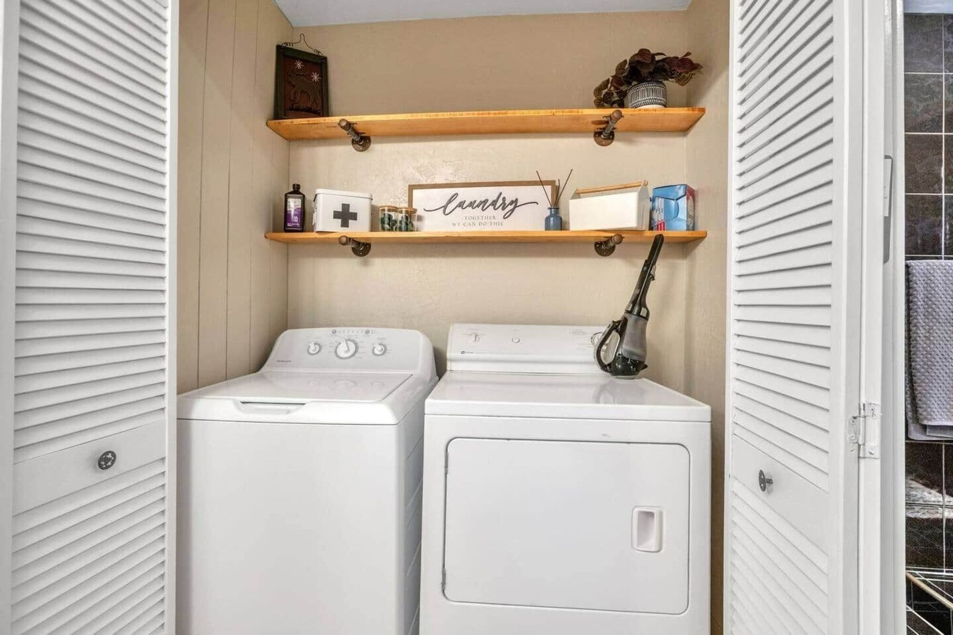 Laundry closet with a white washer and dryer, wooden shelves, and folded decor.