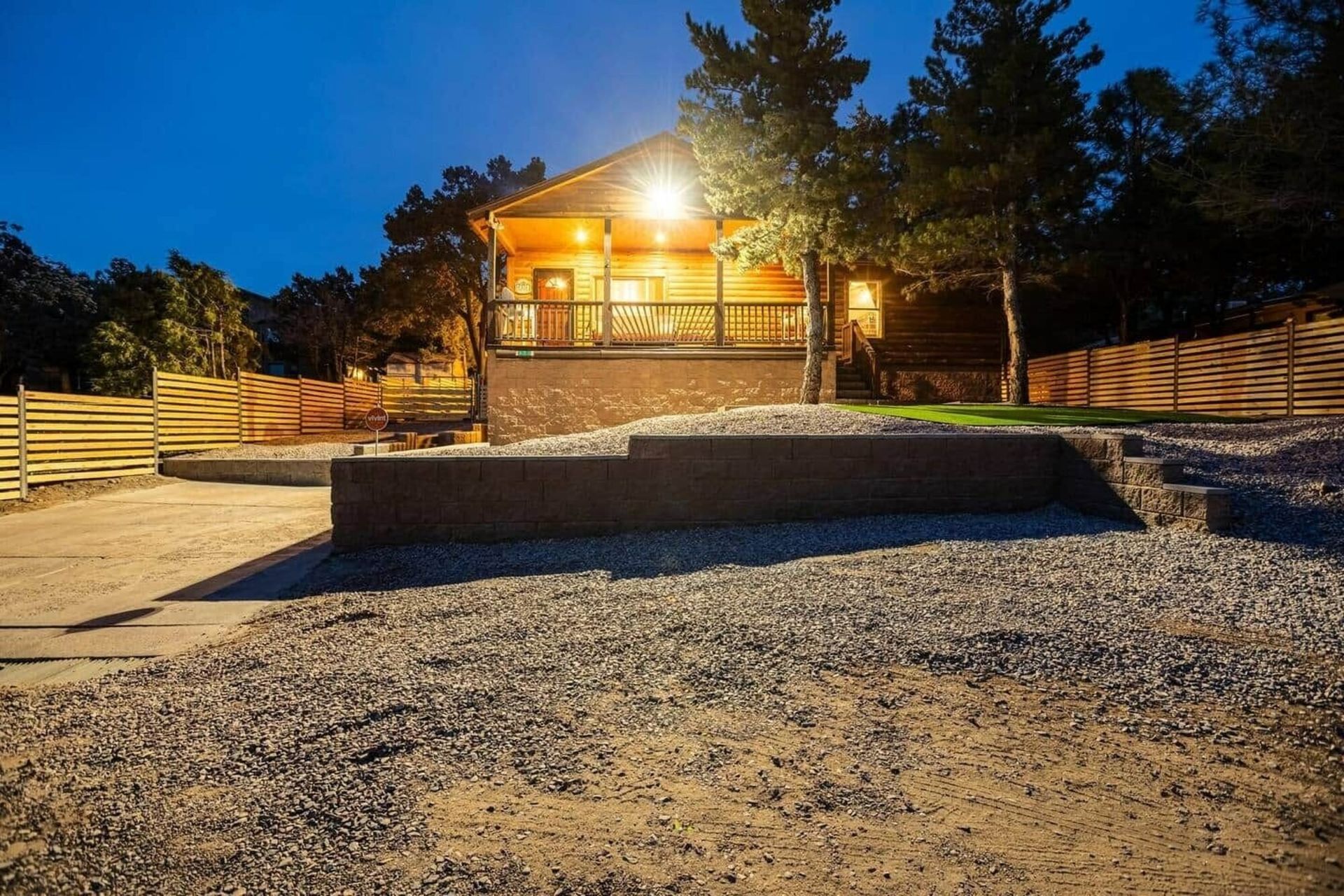 Lit wooden cabin at dusk behind a stone wall, with gravel foreground and trees silhouetted against a blue sky