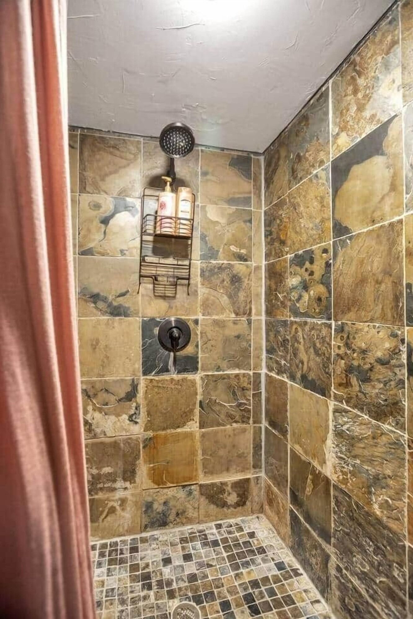 Small tiled shower stall with stone-look walls, mosaic floor, showerhead, and a hanging shower caddy.
