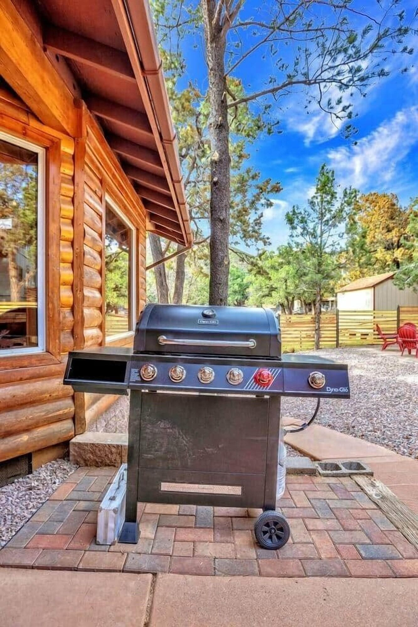 Gas grill on a brick patio beside a log cabin, with trees and a blue sky in the background