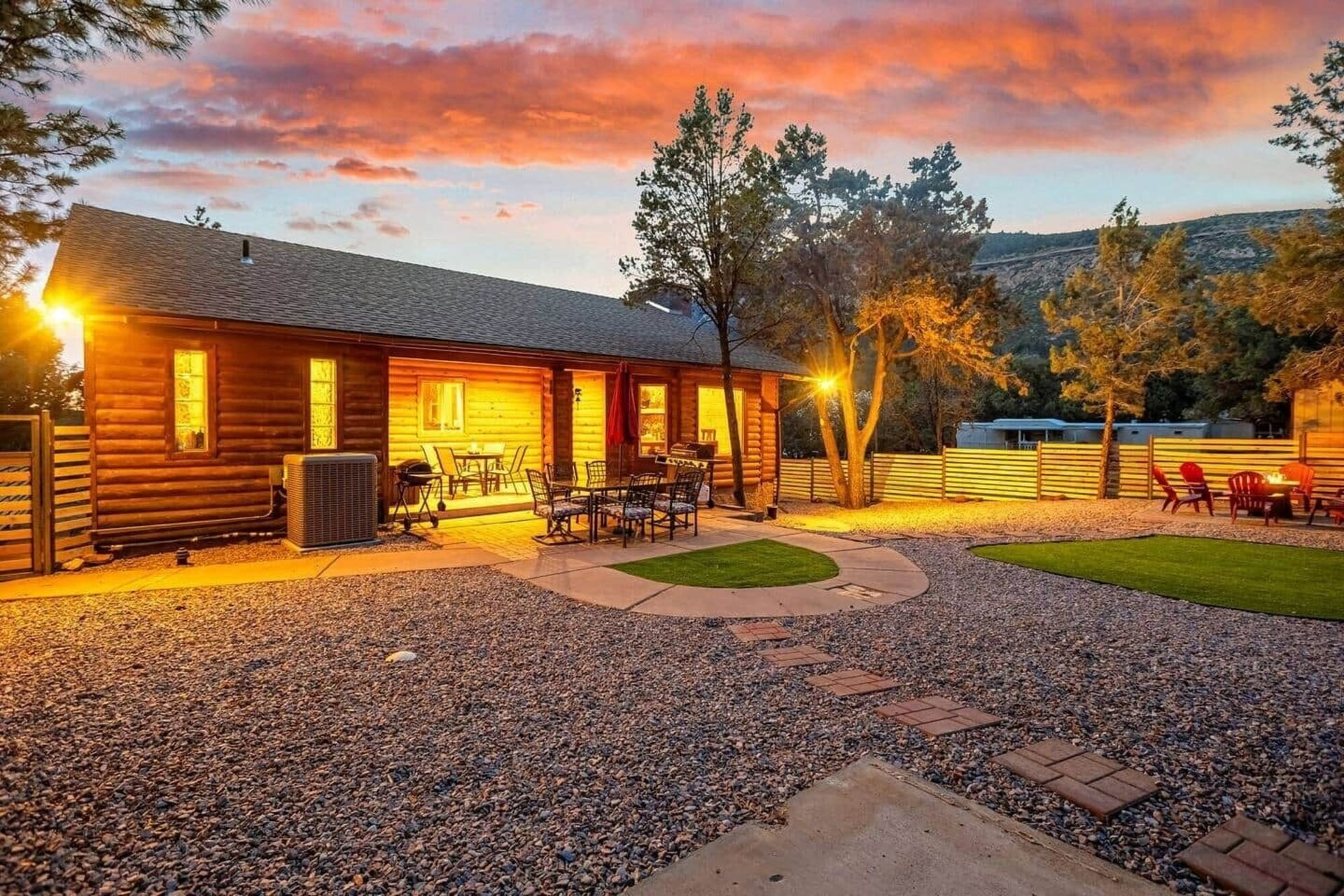 Cozy log cabin at sunset with warm lights, gravel yard, and patio seating under a colorful sky