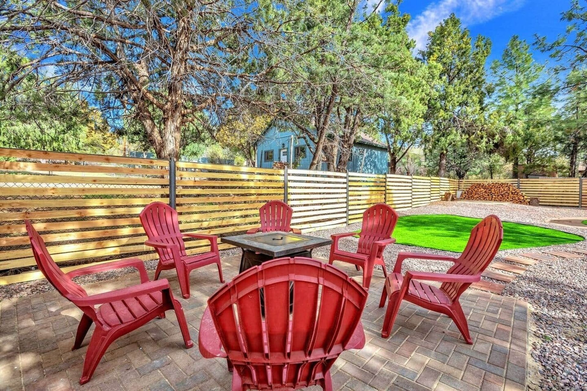 Red Adirondack chairs вокруг круглого костра на каменном патио во дворе под деревьями и голубым небом