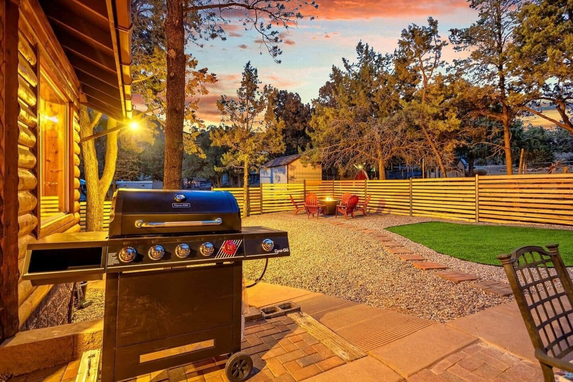 Patio with gas grill, wooden deck, stone path, green lawn, and sunset-lit backyard fence