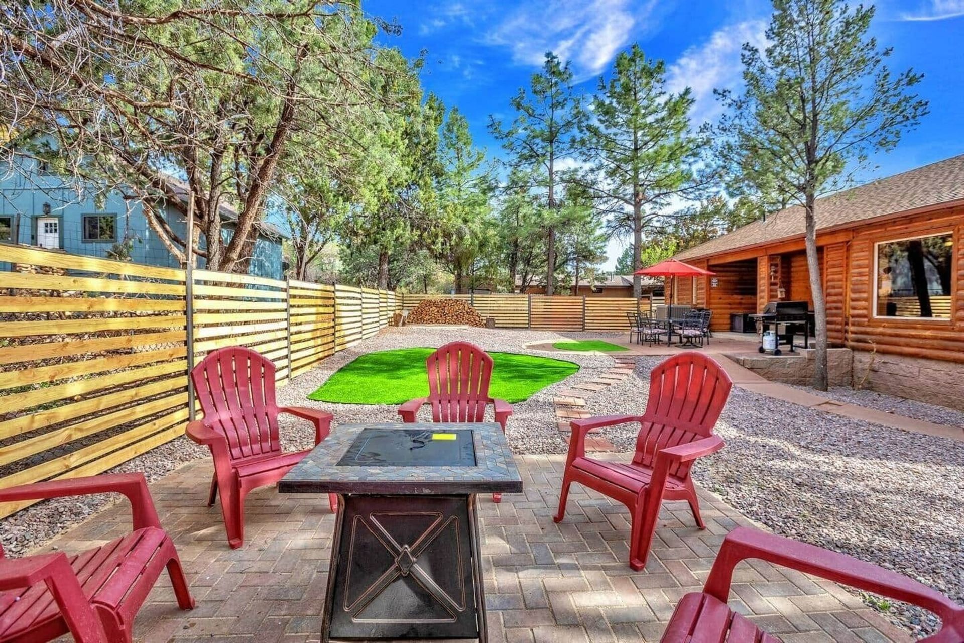Sunny backyard patio with red chairs, fire pit table, and wooden fence beside a house