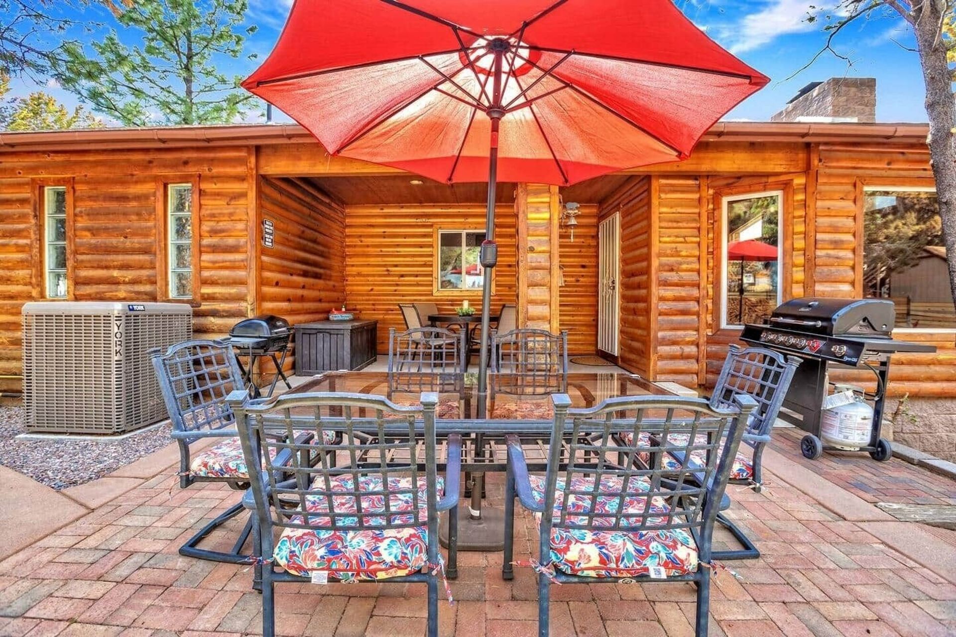 Outdoor log cabin patio with red umbrella, metal chairs, and grill on a brick terrace