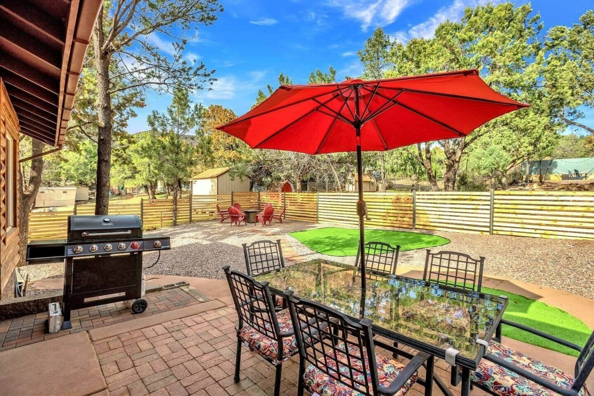 Patio with red umbrella, glass table, metal chairs, and a grill in a landscaped backyard
