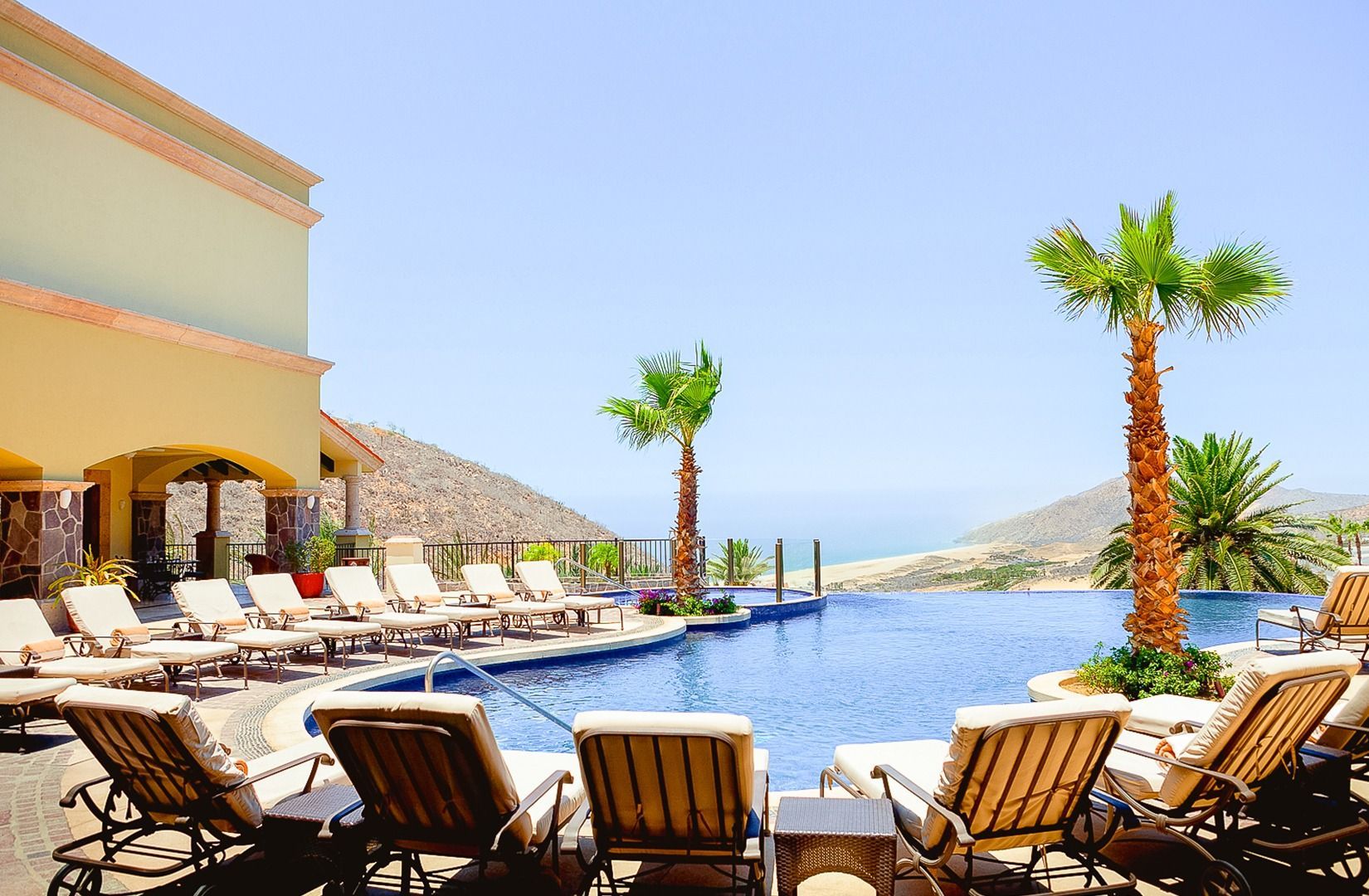 Poolside lounge chairs beside a resort infinity pool with palm trees and a bright blue sky