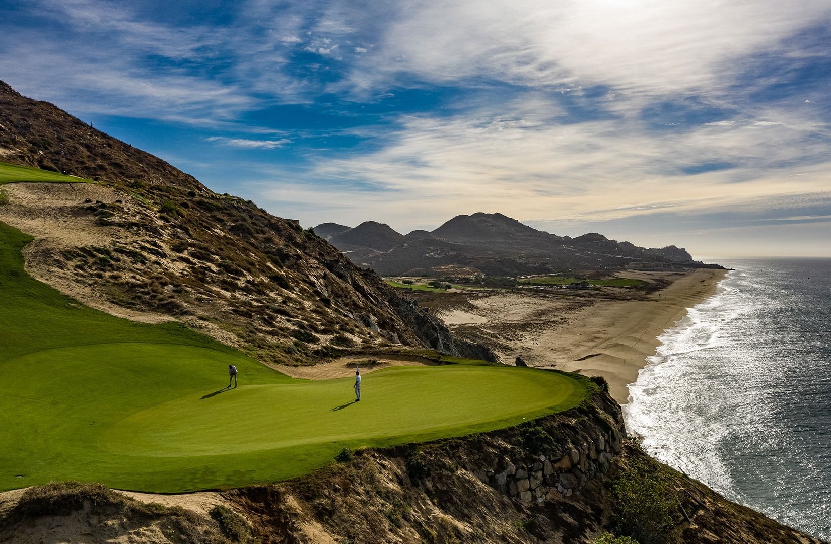 Green golf course on a cliff overlooking the ocean, with rocky hills and a cloudy sky.