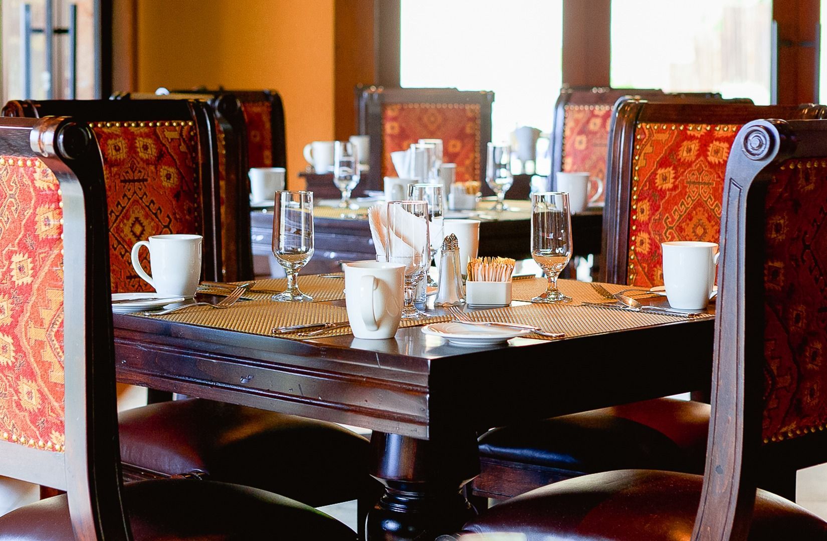 Dining table set with white cups, glassware, and red patterned chairs in a warm restaurant interior