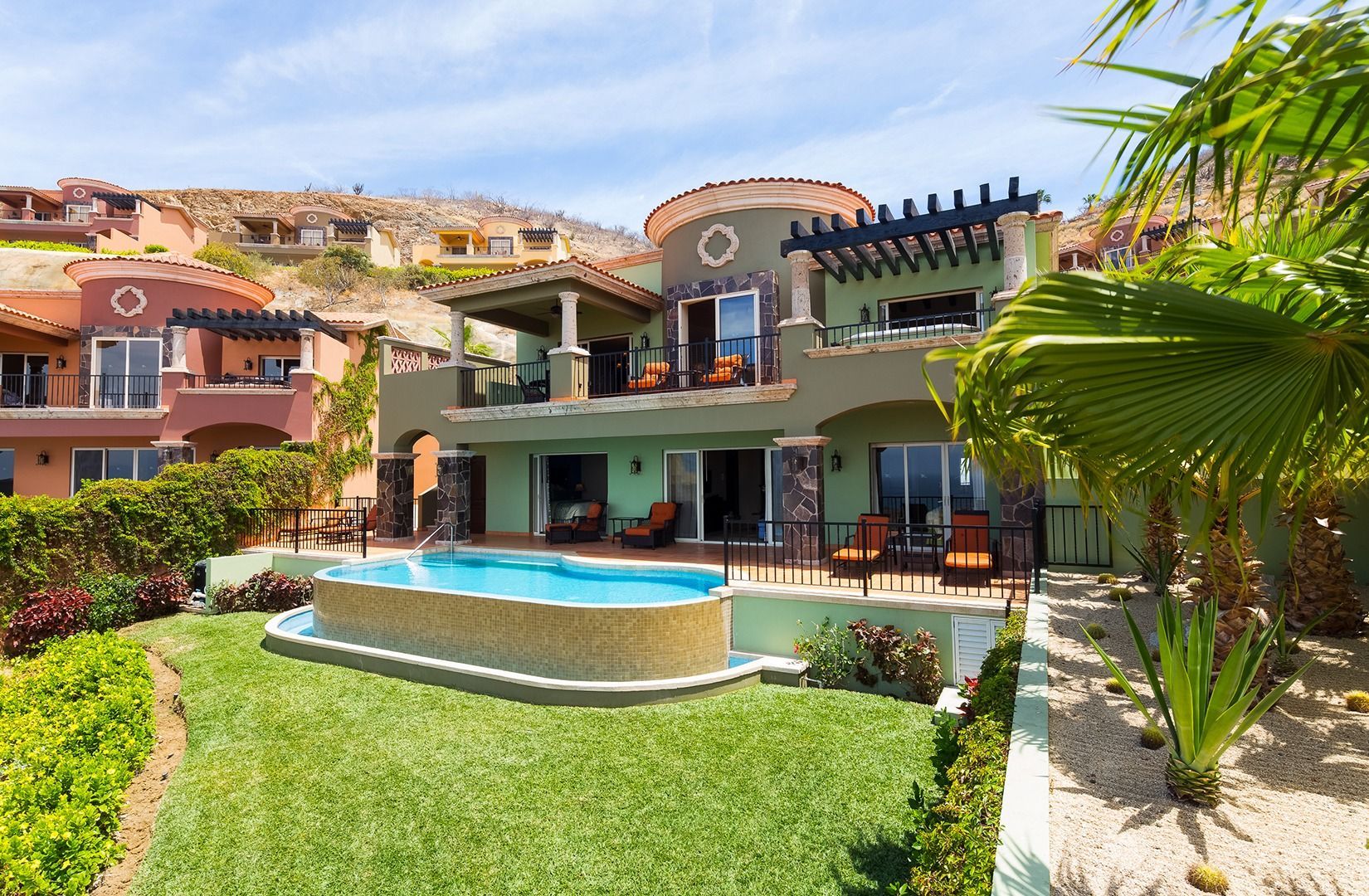 Modern villa with private pool, green lawn, and palm trees in a sunny hillside resort setting
