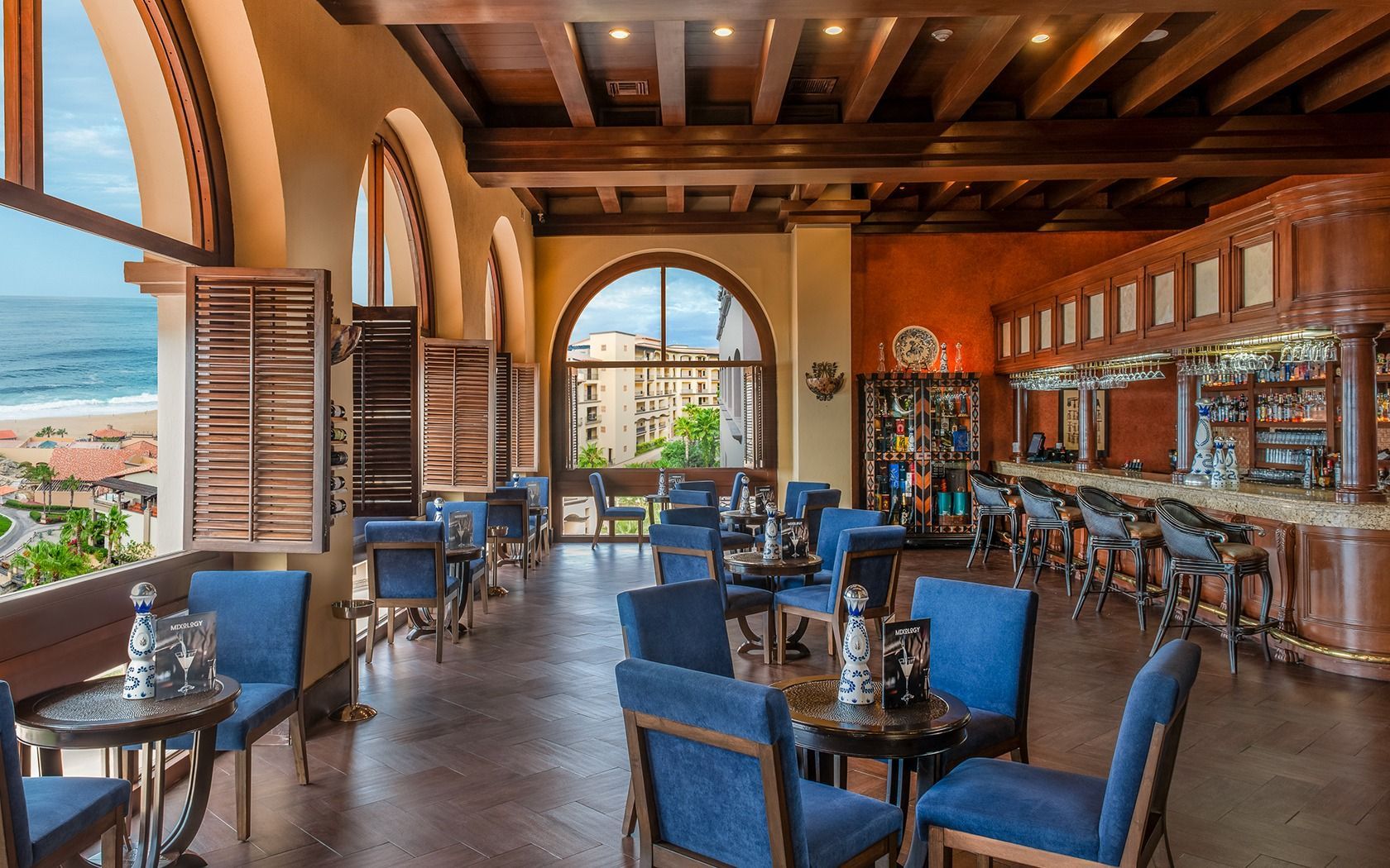 Elegant restaurant interior with blue chairs, arched windows, wood beams, and ocean views