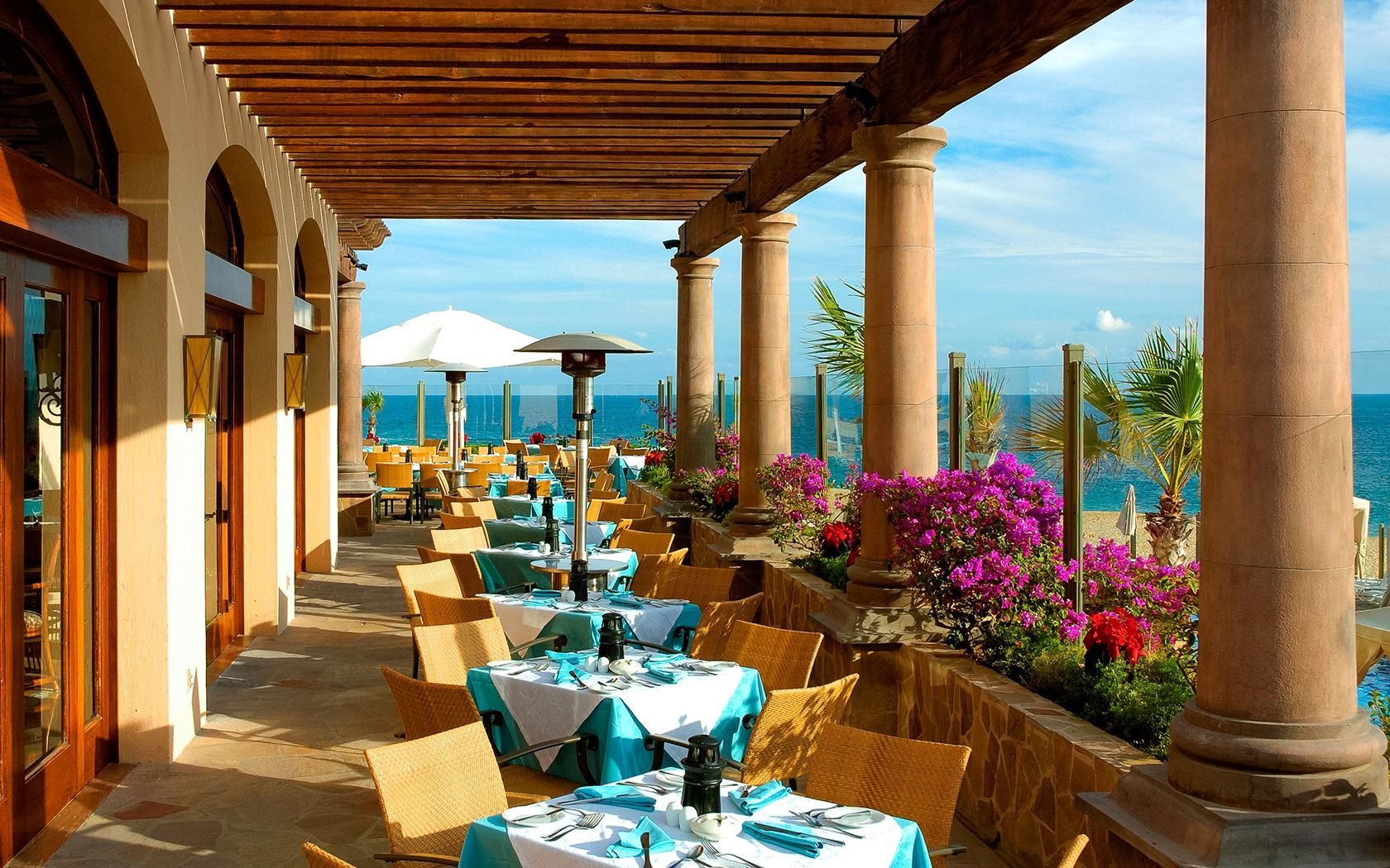 Seaside restaurant patio with turquoise tables, wicker chairs, columns, and ocean views
