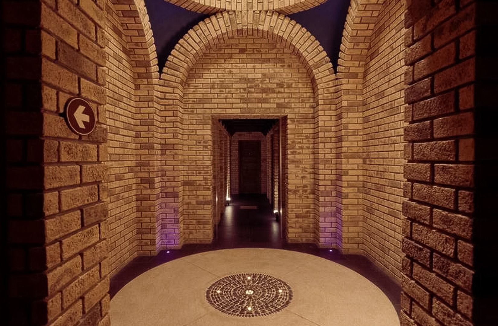 Arched brick corridor with warm lighting, tiled floor, and a dark doorway at the end.