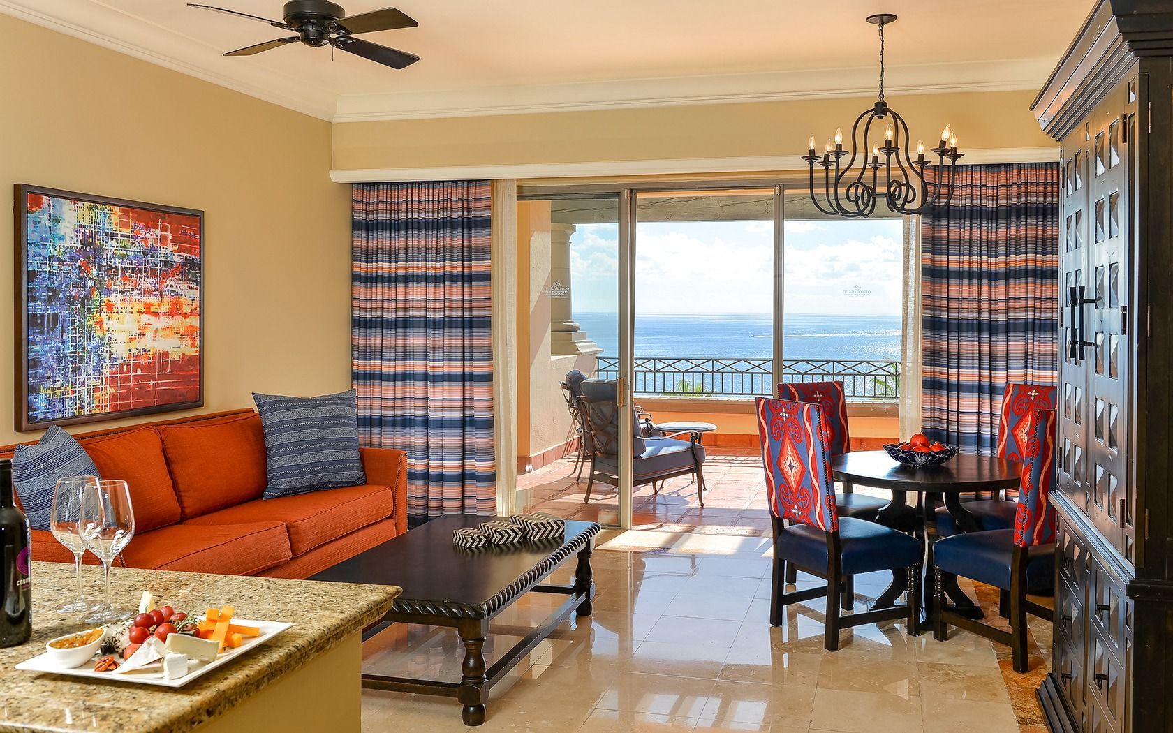 Bright living-dining room with orange sofa, table, chandelier, and ocean-view balcony.