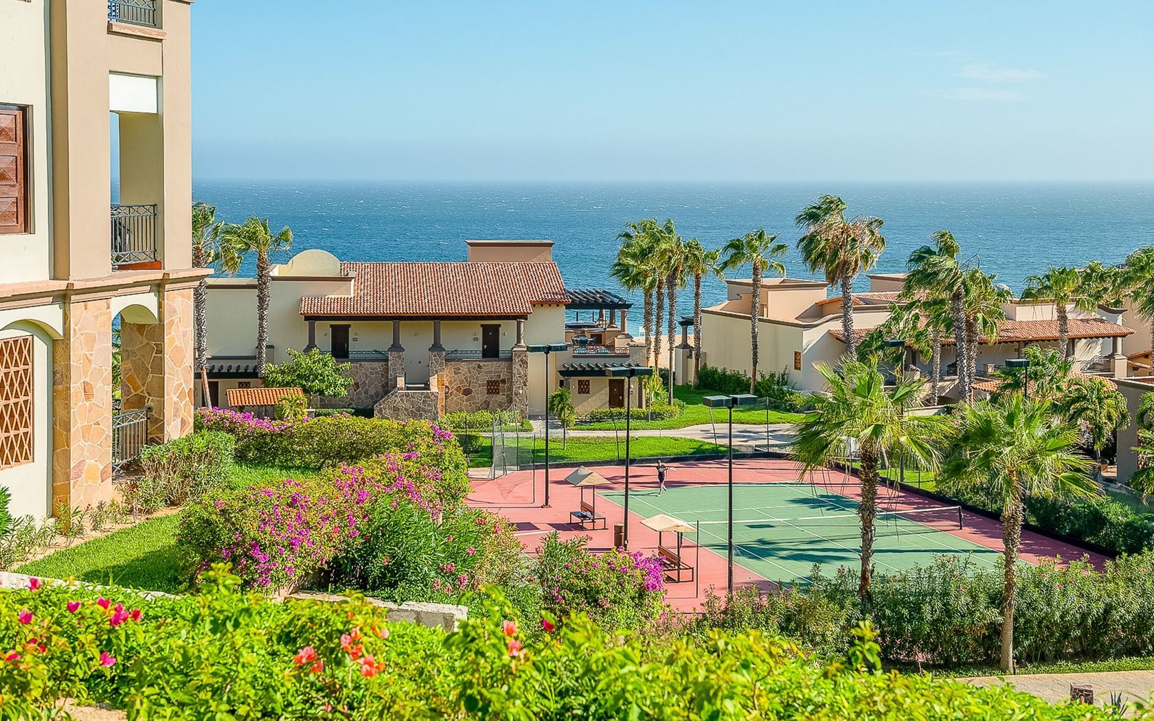 Resort courtyard with pink tennis court, palm trees, and ocean view under a clear blue sky