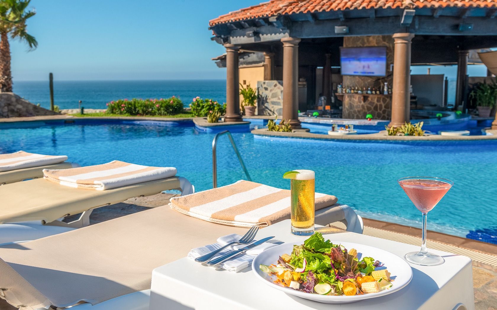 Poolside resort patio with drinks and salad overlooking a blue ocean and palm trees
