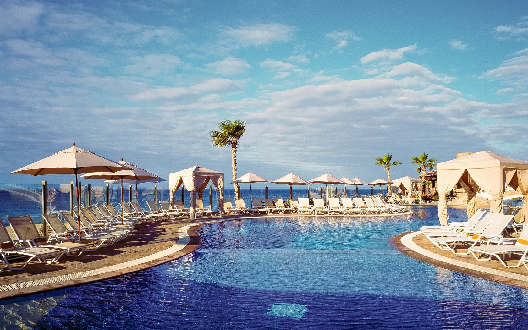 Luxury resort pool with blue water, white lounge chairs, cabanas, and ocean views under a sunny sky