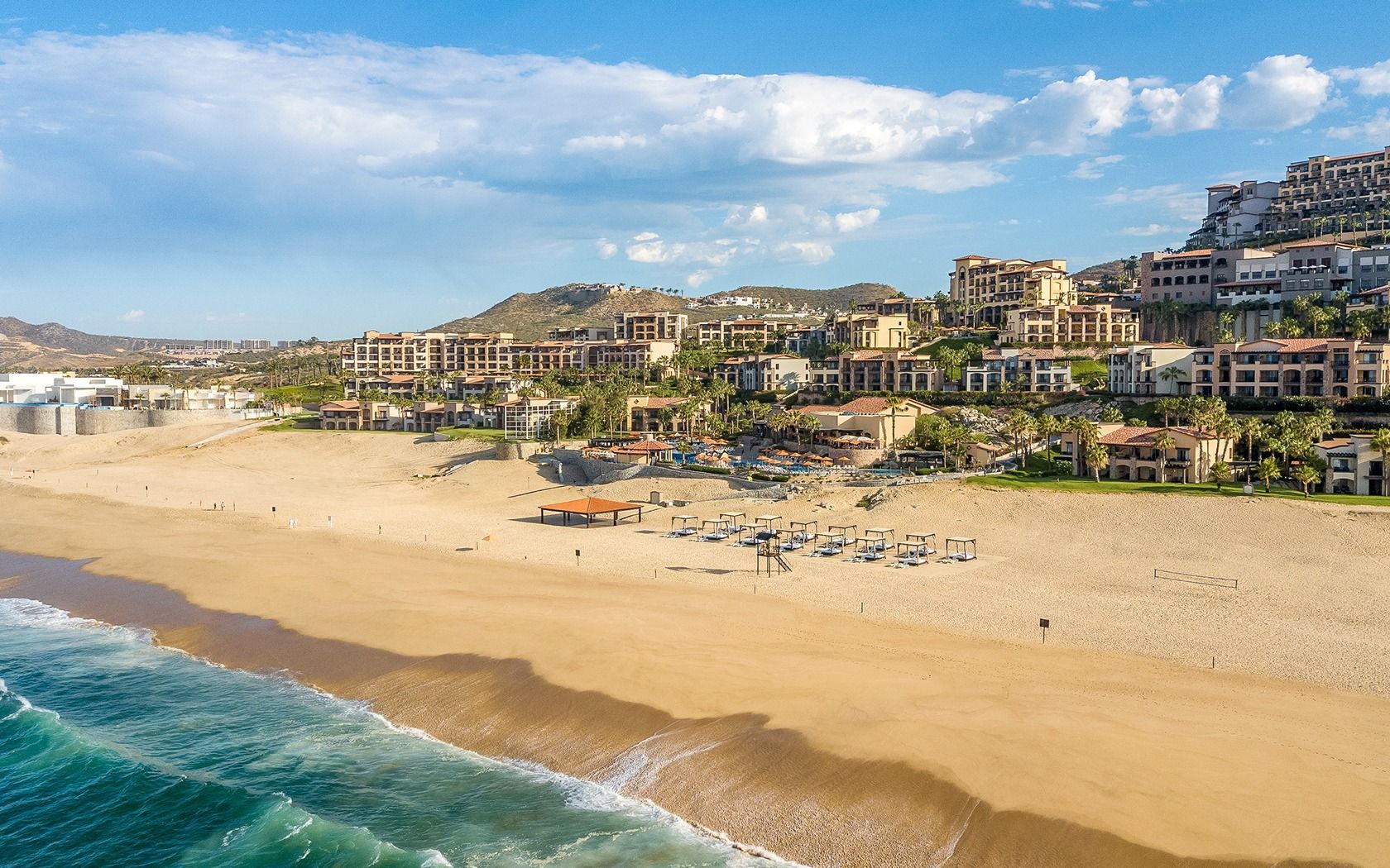 Sunny beach with turquoise waves, wide sandy shore, and hillside resort buildings under a blue sky