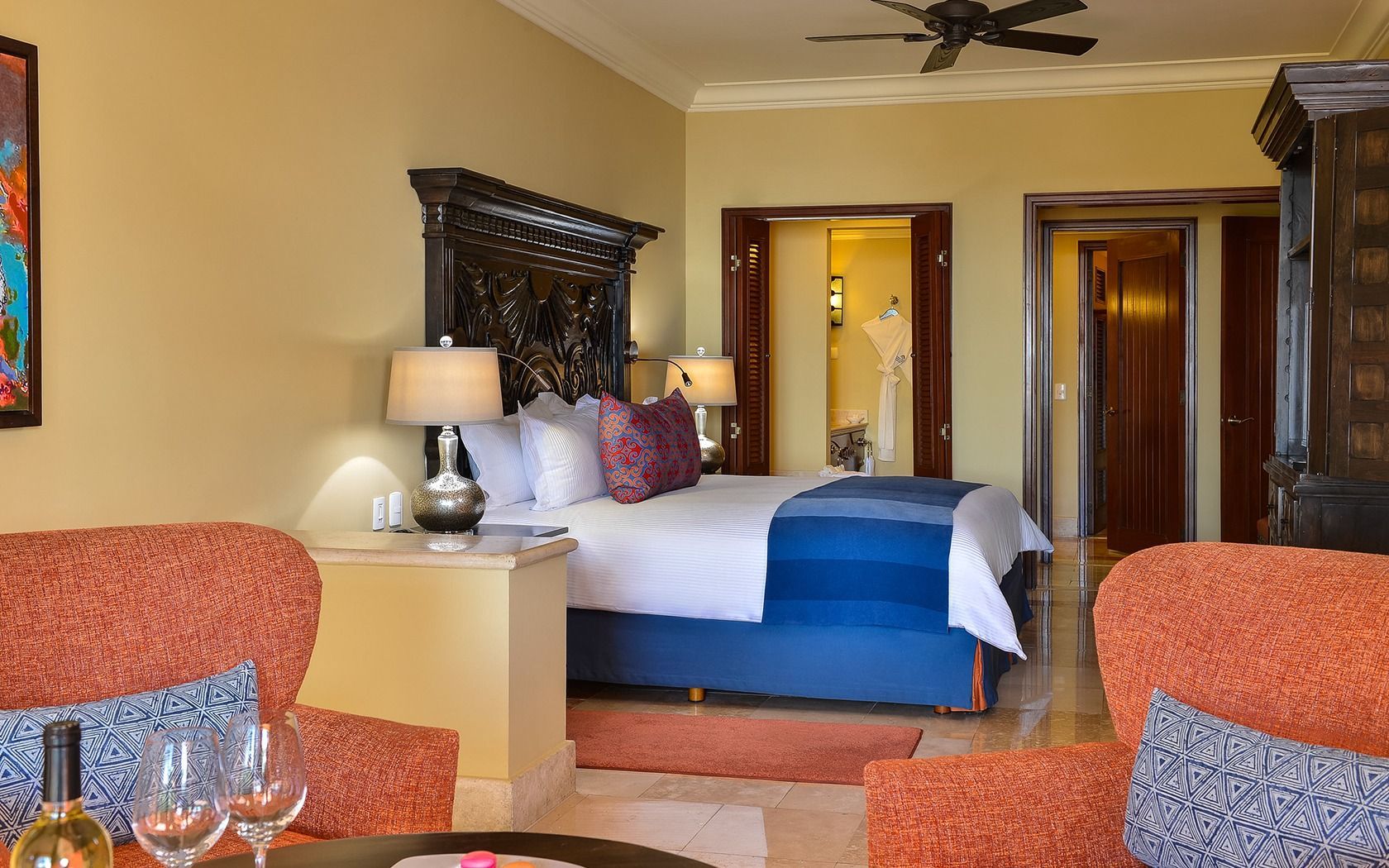Hotel bedroom with blue bed, ornate headboard, lamp, and orange armchairs in a warm-toned room