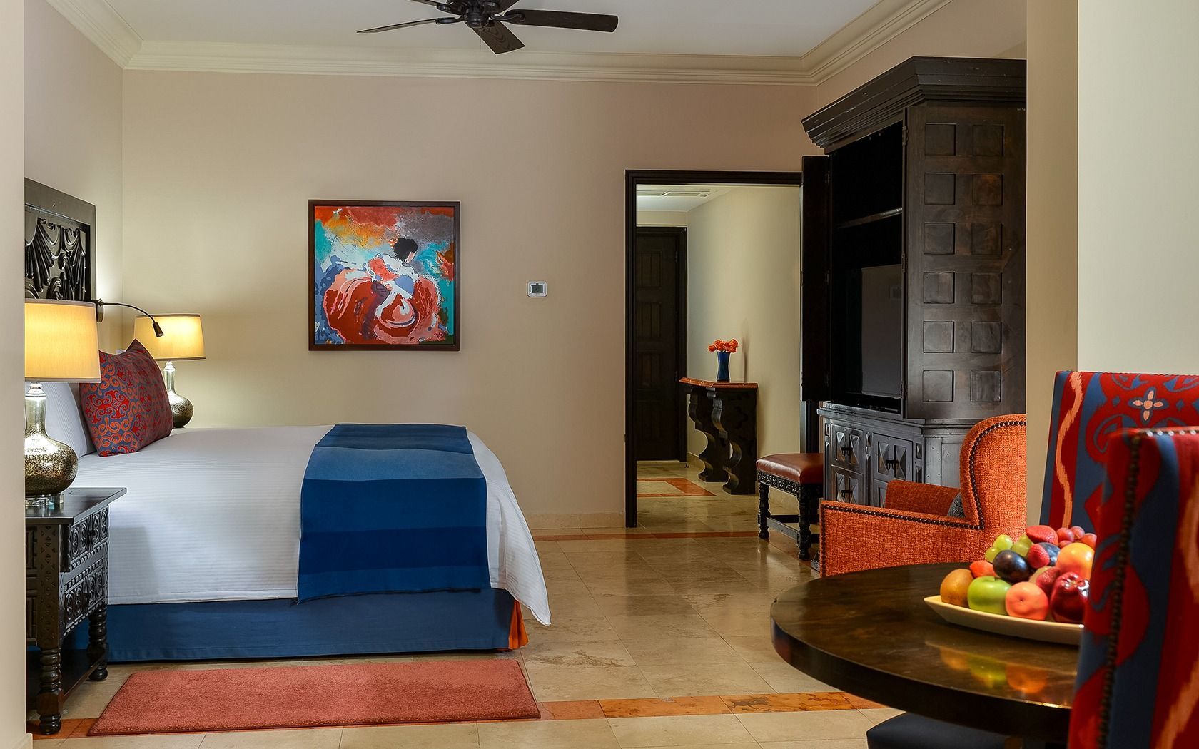 Modern bedroom with blue bedspread, colorful art, warm lamps, and a wooden dresser.