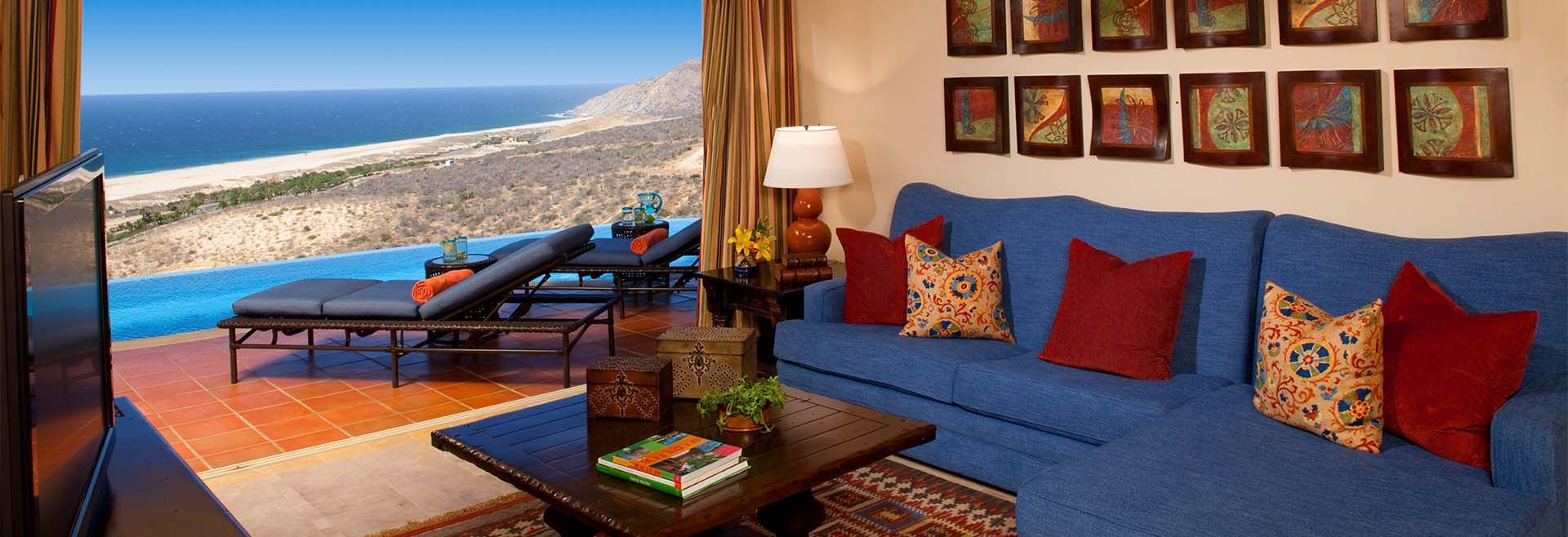 Ocean-view living room with blue sofa, wicker chairs, and colorful pillows overlooking a beach and sea through open doors