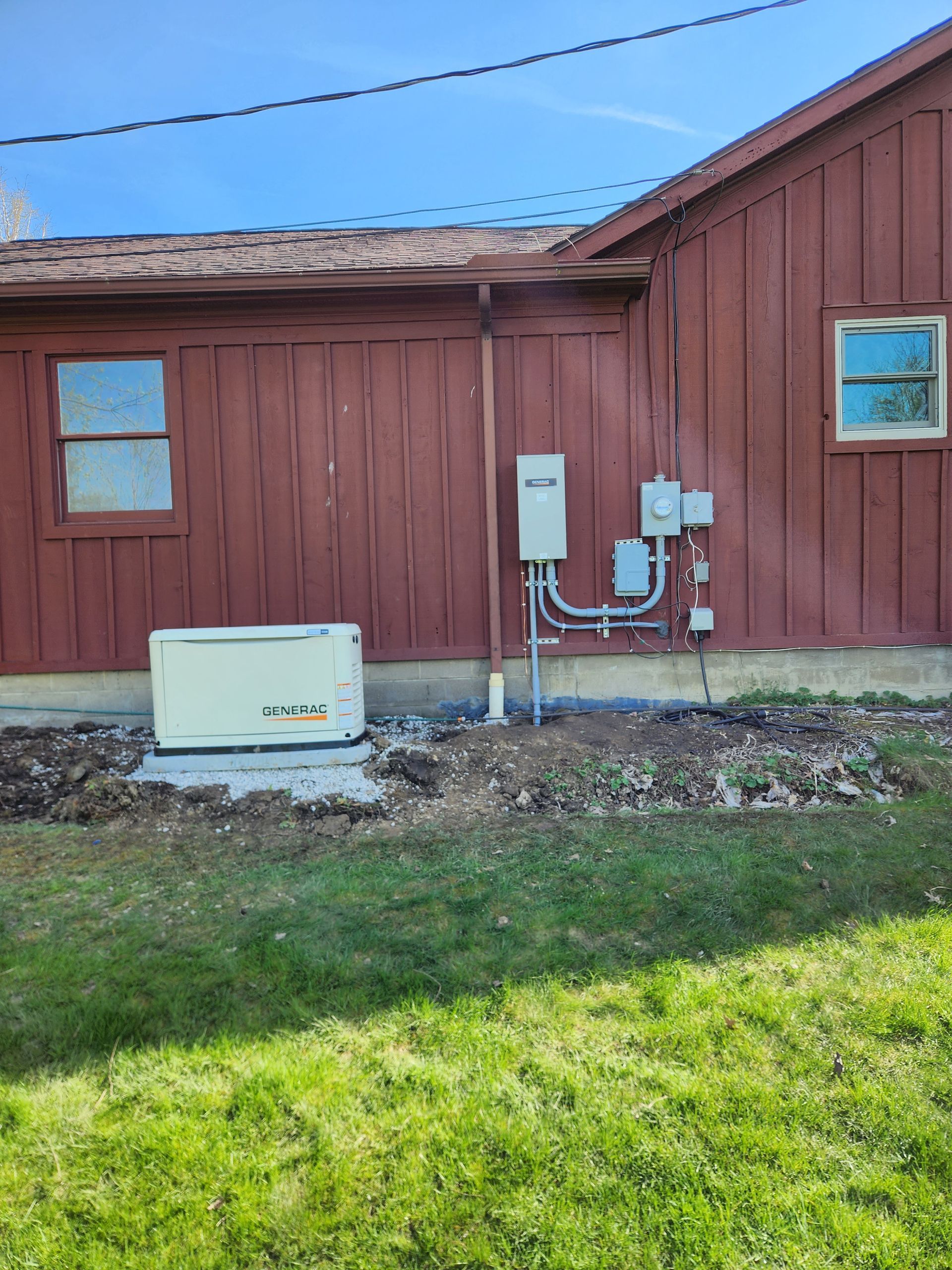 A generator installed next to a red-painted building with electrical components and windows. Green grass in the foreground.