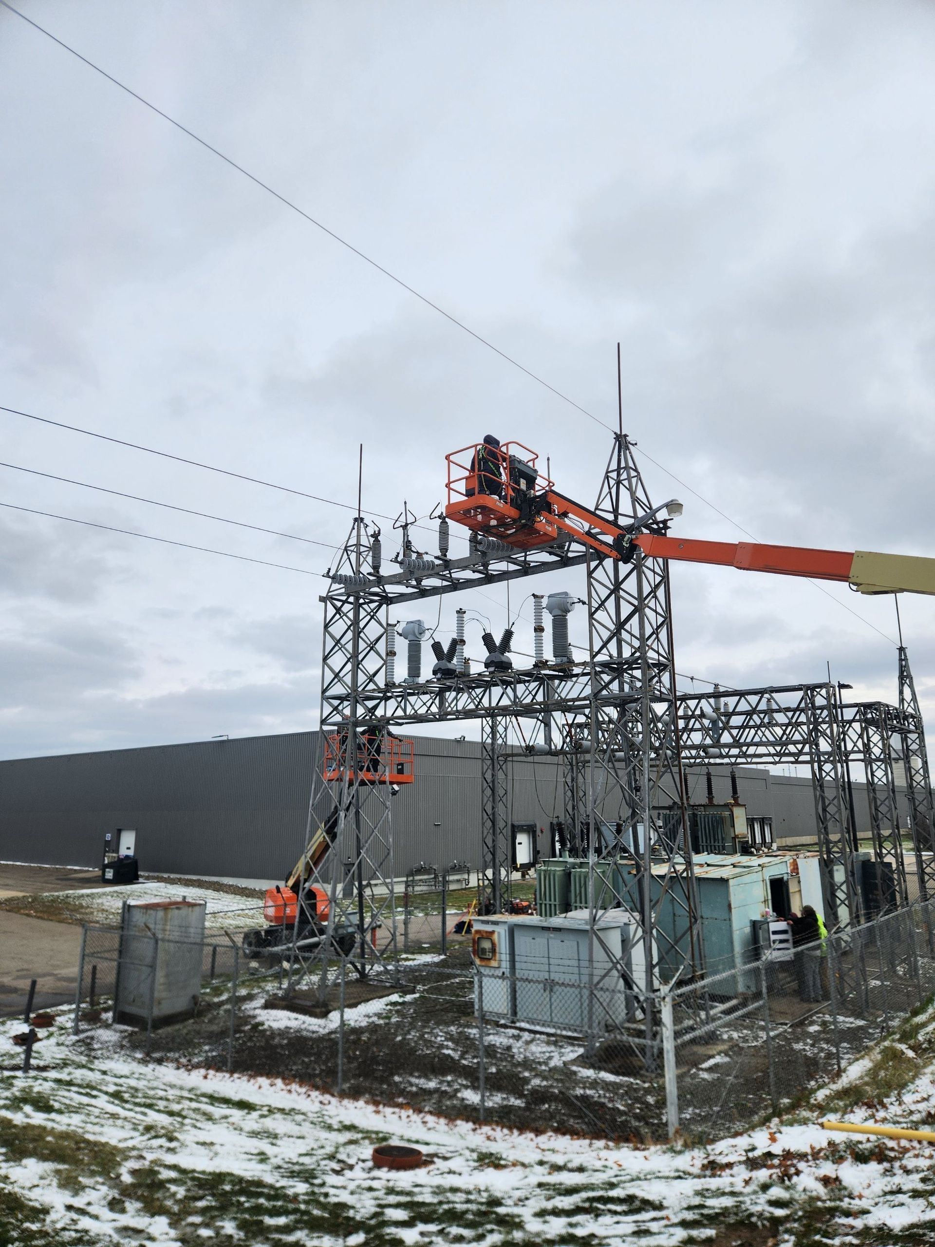 Workers in orange lifts at an electrical substation on a cloudy day, snow on the ground.