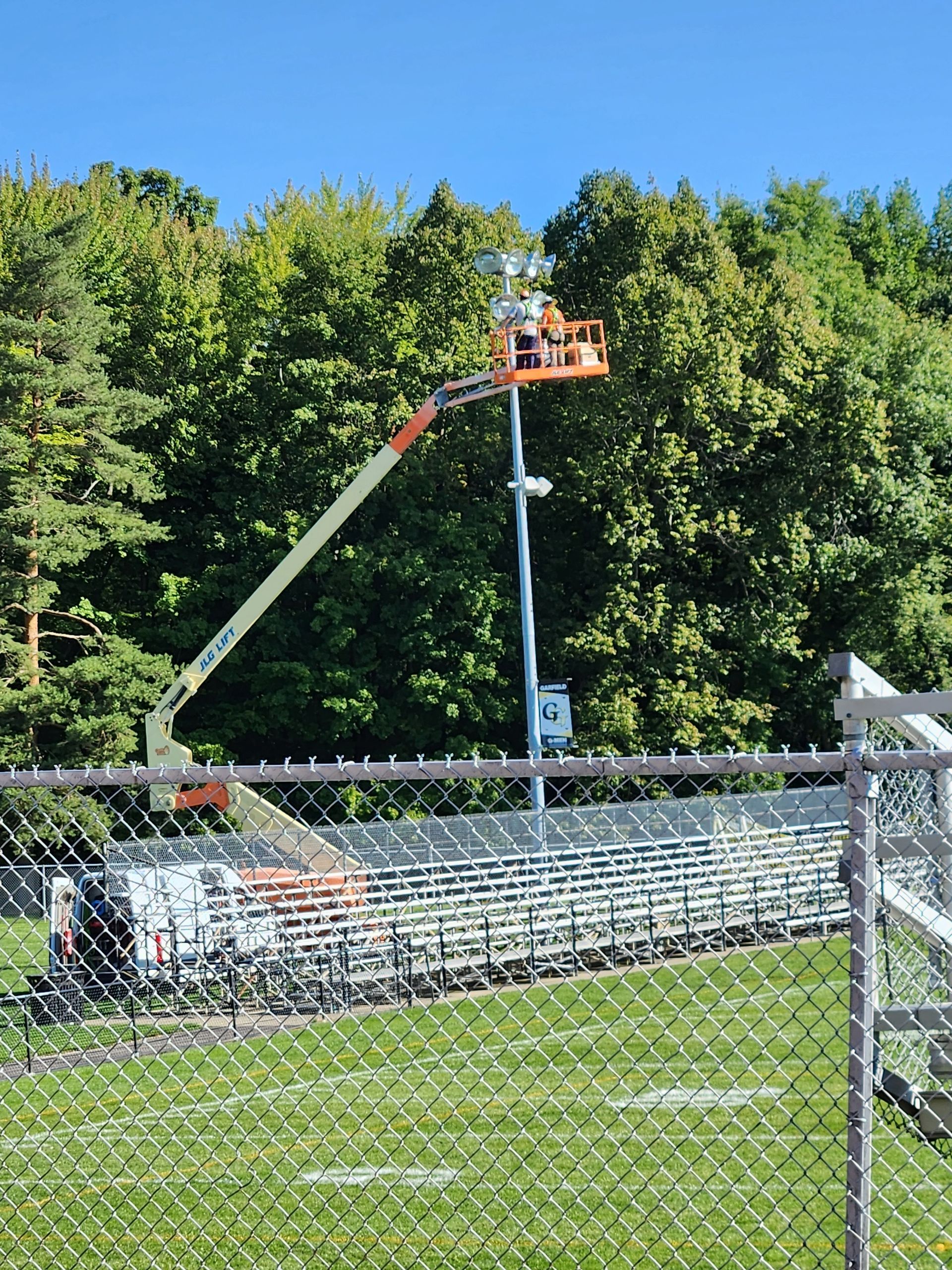 A person in a lift working on a light pole above a stadium, near a forest.