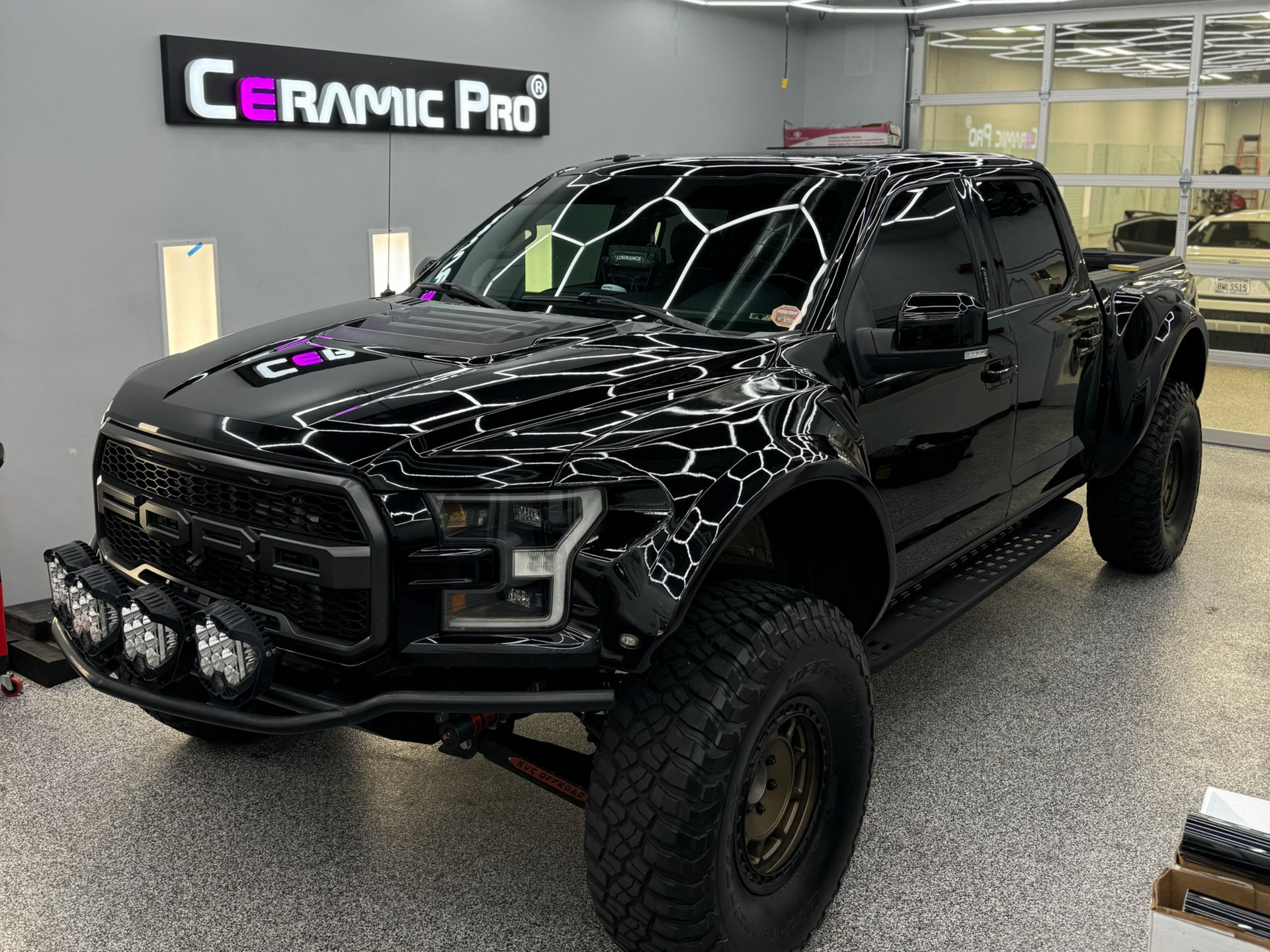 A black Ford Raptor truck parked in a well-lit workshop with a Ceramic Pro sign on the wall.