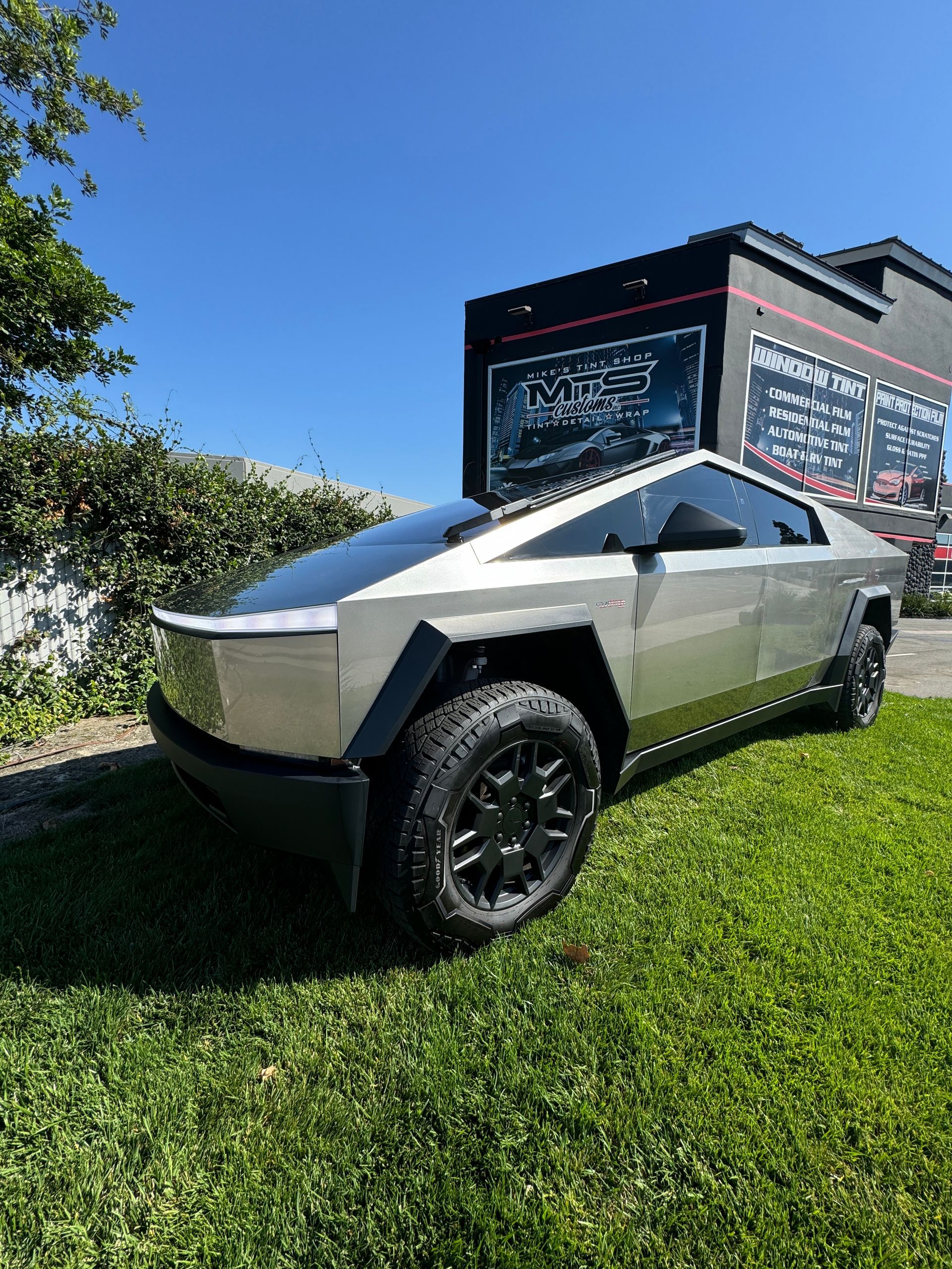 Tesla Cybertruck parked on grass with a black building in the background. Sunny day.