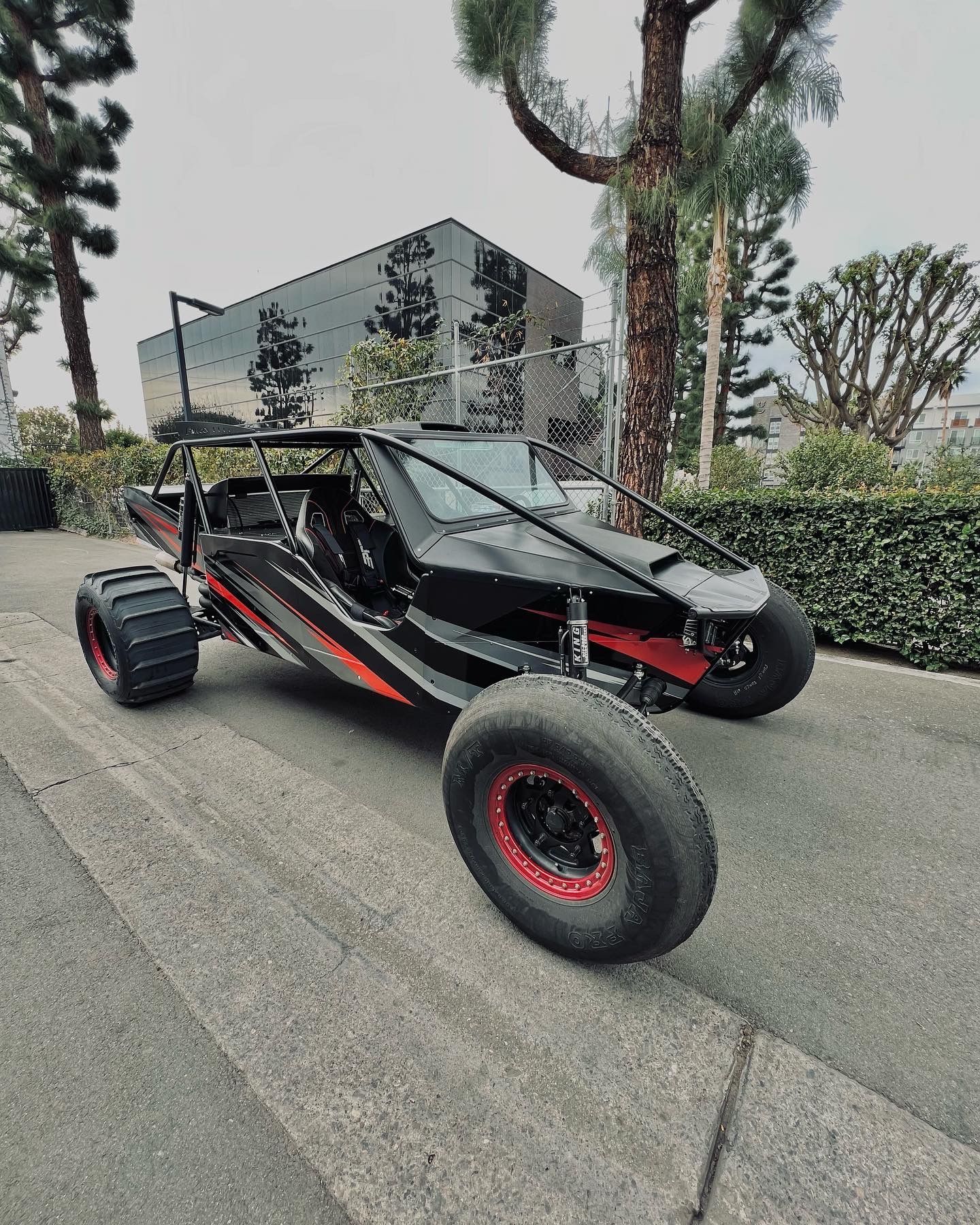 Black off-road dune buggy with red accents parked on a street beside hedges and a modern building.