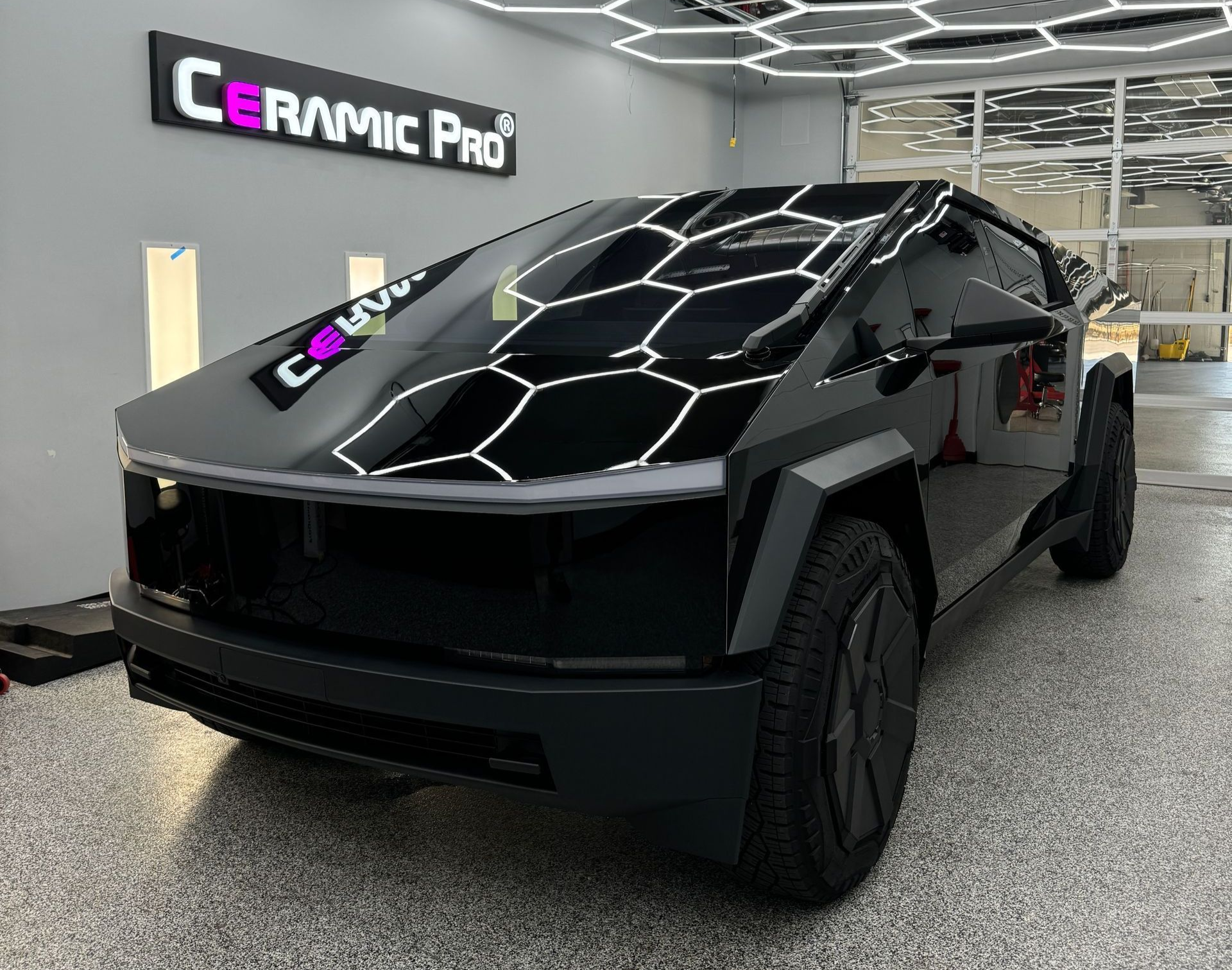 A black Tesla Cybertruck parked in a garage with hexagonal ceiling lights reflecting on its polished, wrapped exterior.