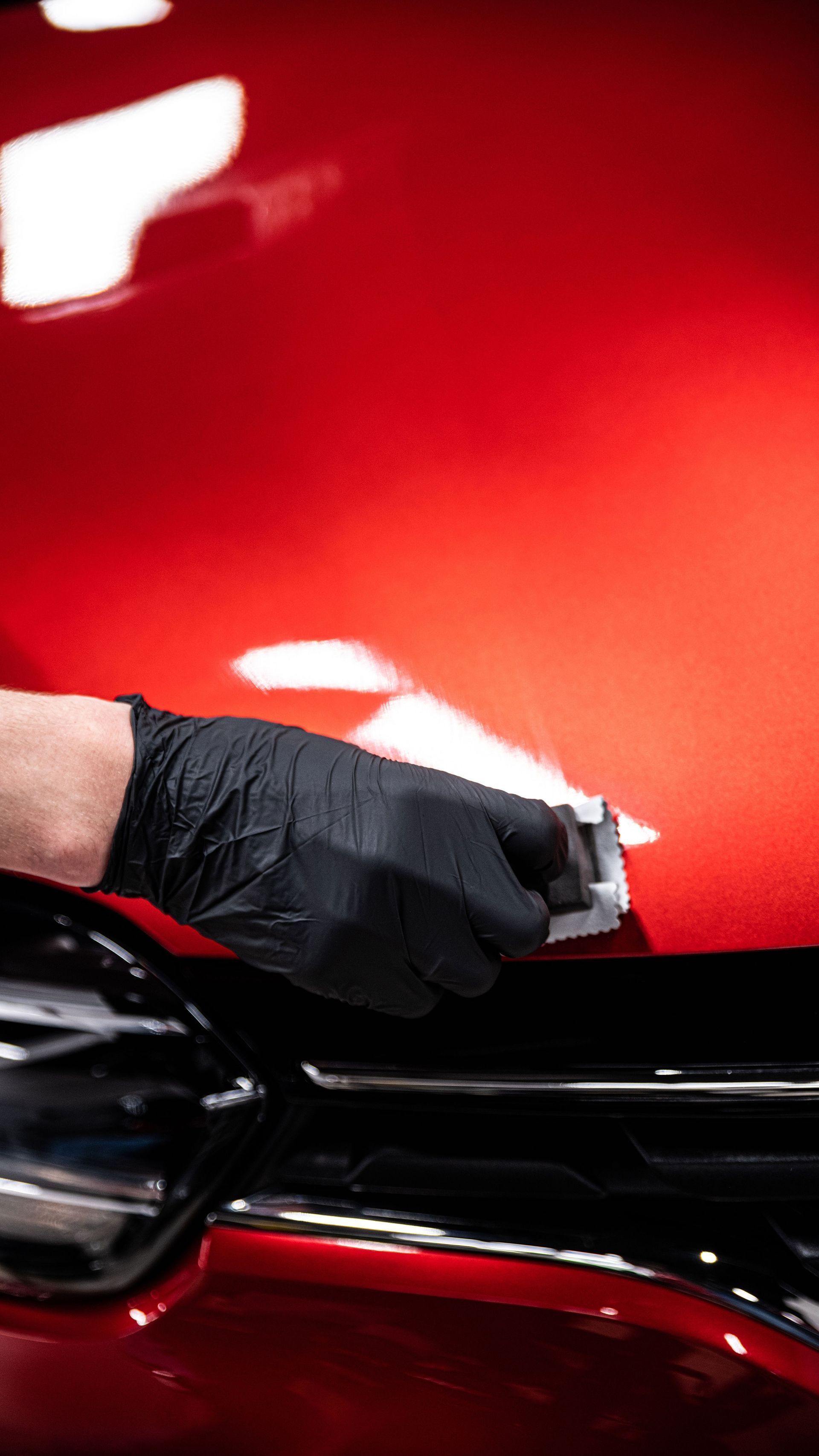 Gloved hand applying a coating to a shiny red car's hood. Black gloves, reflective surface.