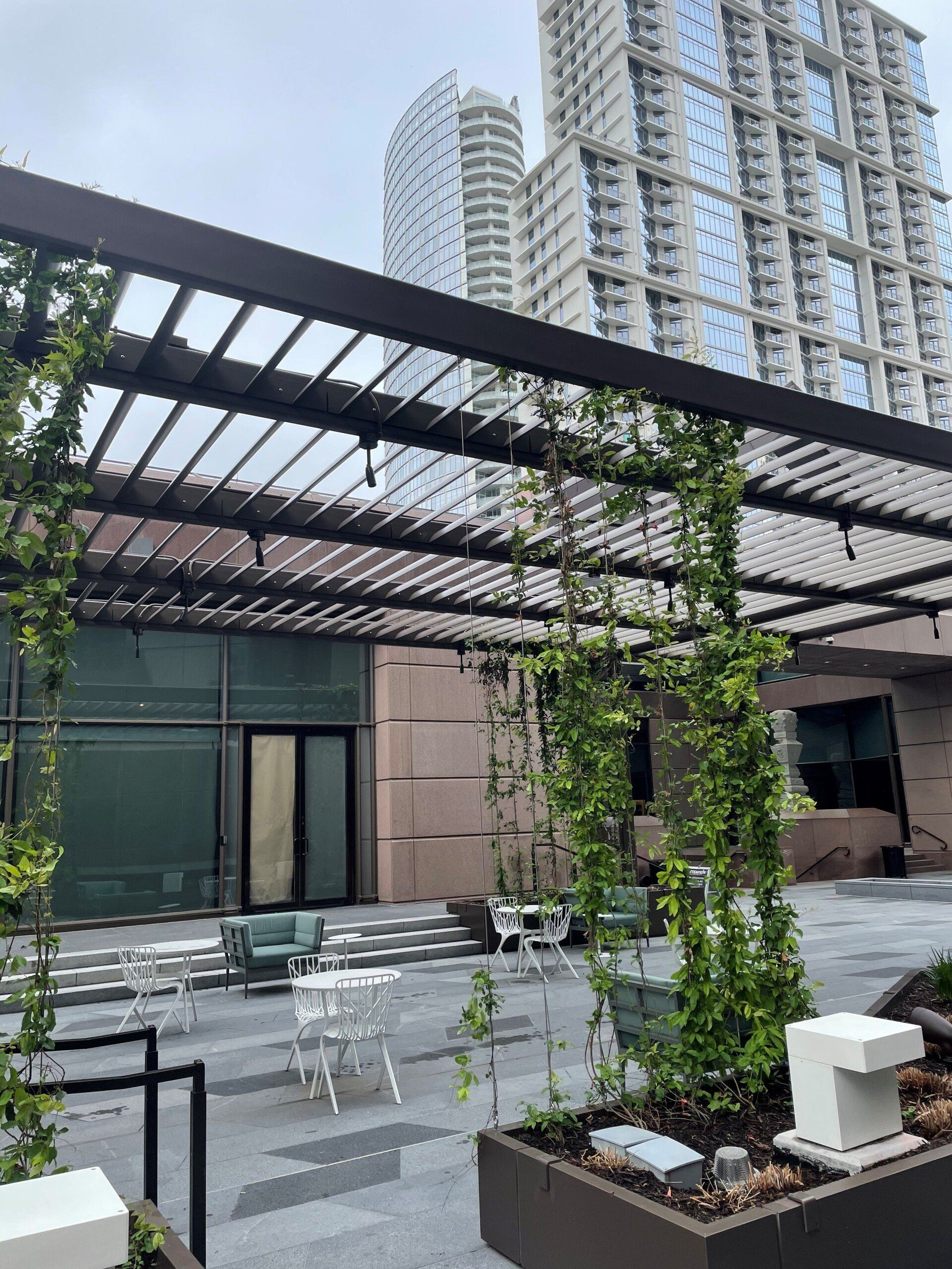 A patio with tables and chairs under a pergola with a building in the background.