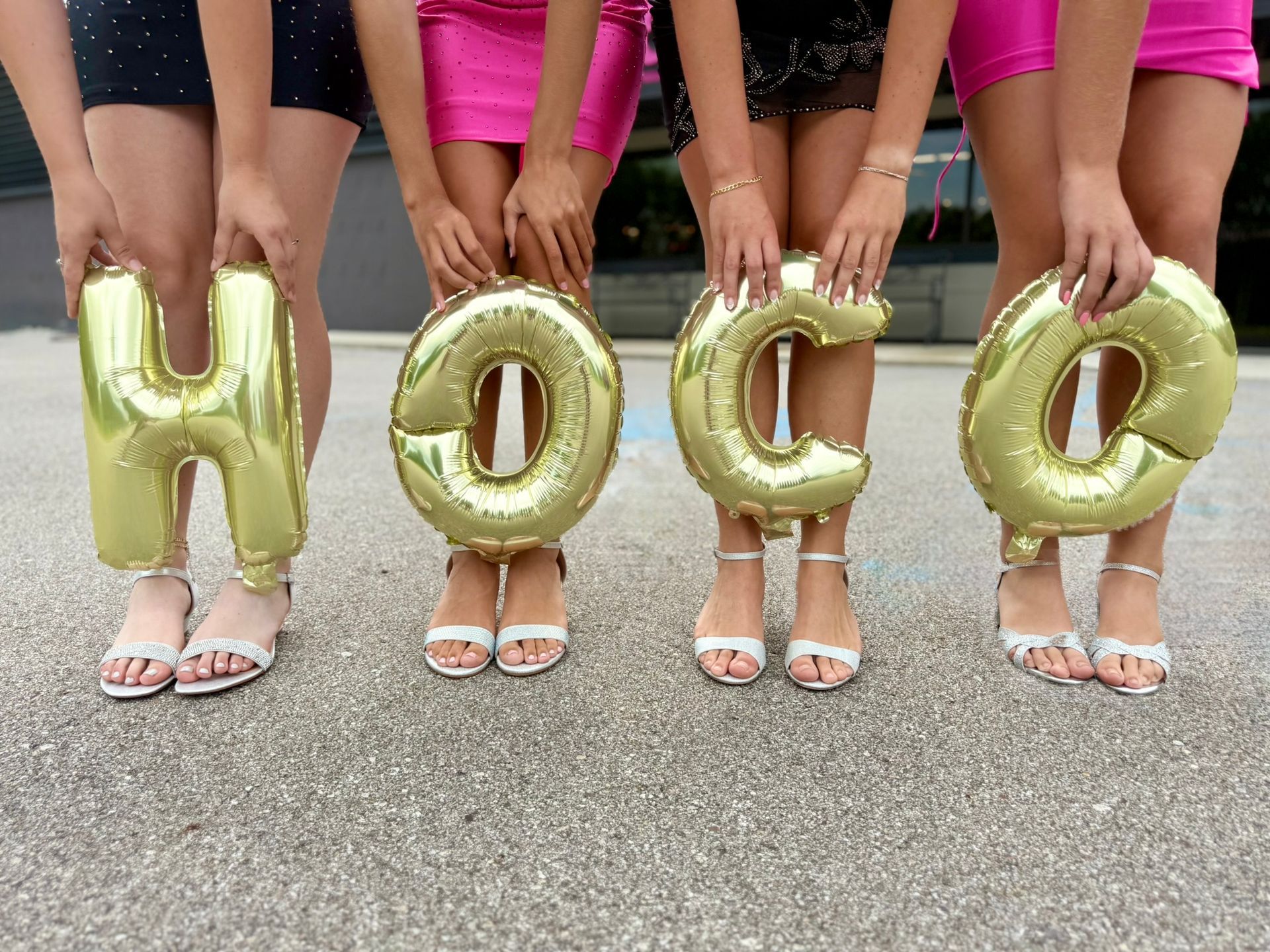 four girls in short formal homecoming dresses in heels holding the letters HOCO