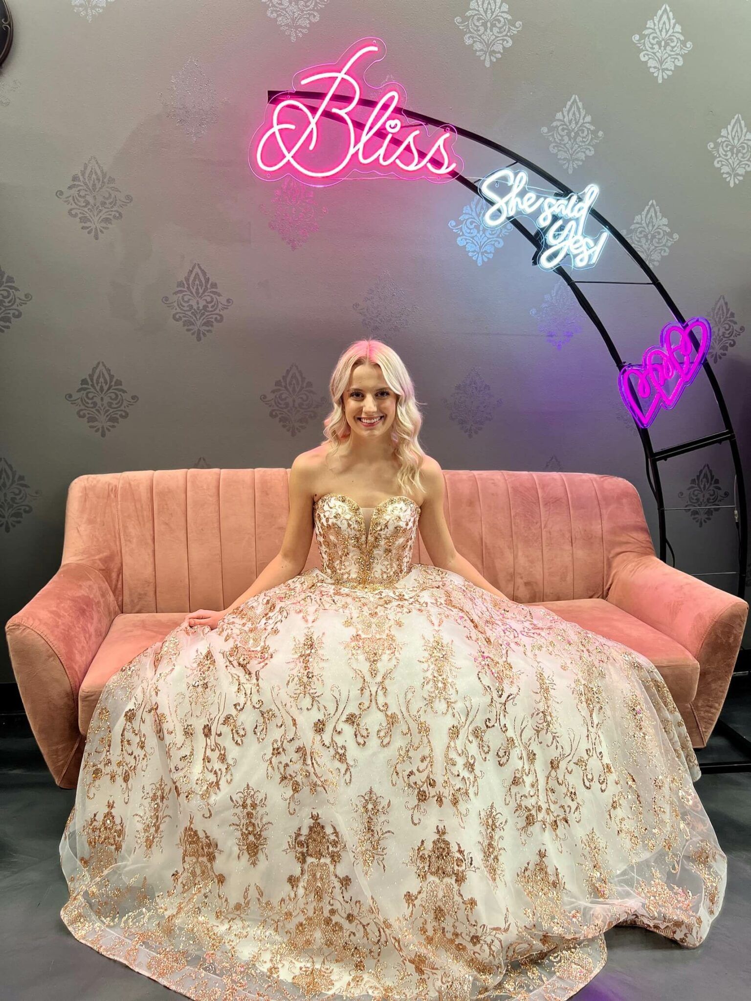 Woman in formal gown, seated on pink couch, under neon sign that says 