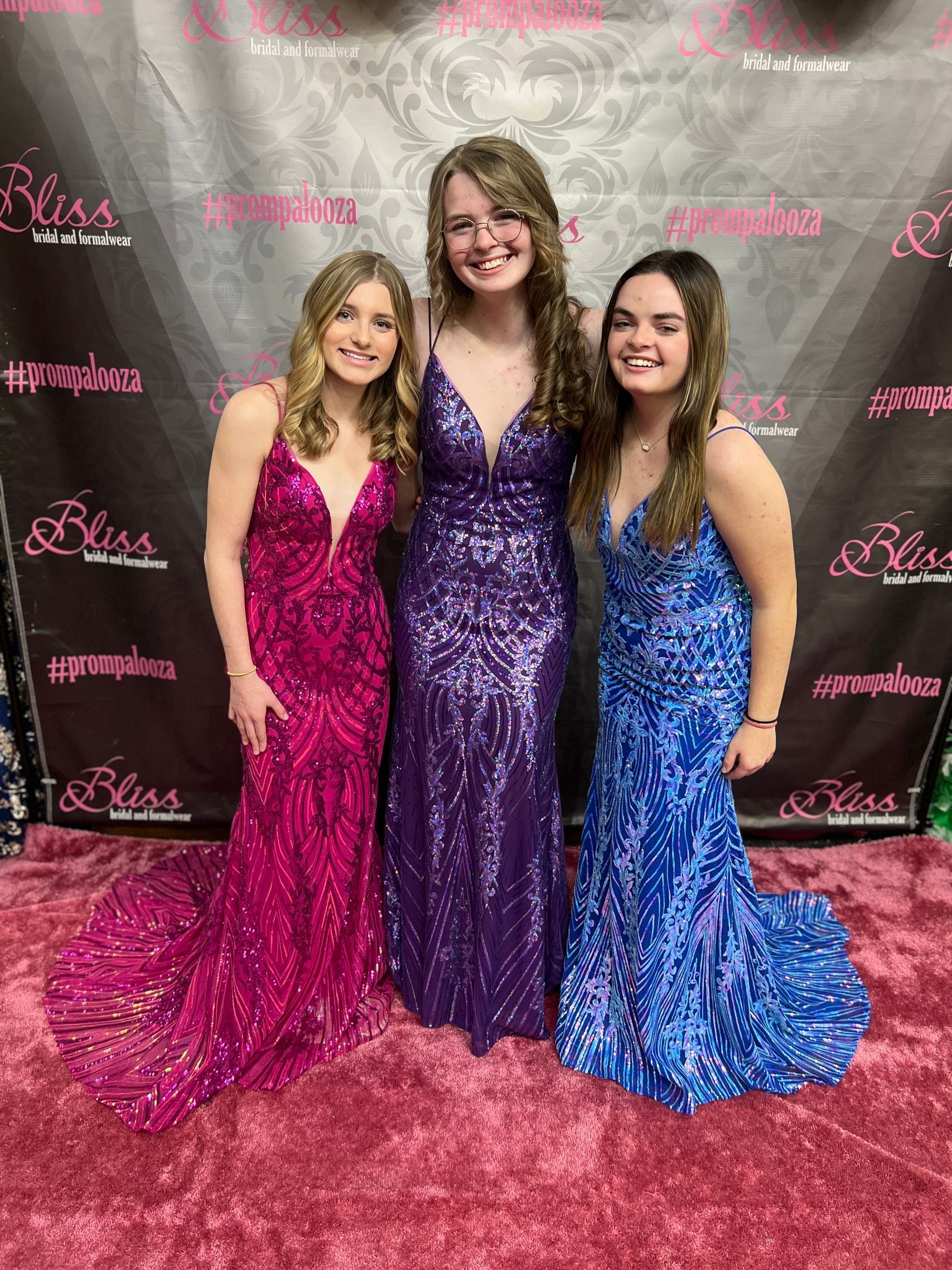 Three young people in formal dresses pose for a photo.  A purple carpet and backdrop.  Dresses are hot pink, purple, and blue.
