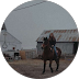 Person on horseback in front of barns on a cloudy day.