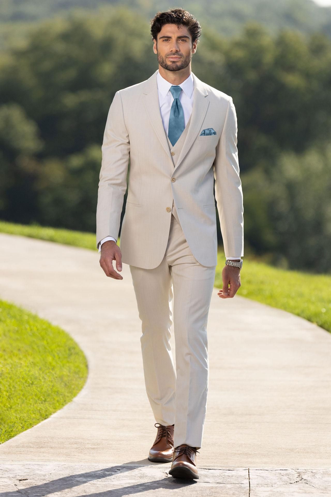 Man in beige suit and teal tie walks on a stone path outdoors.